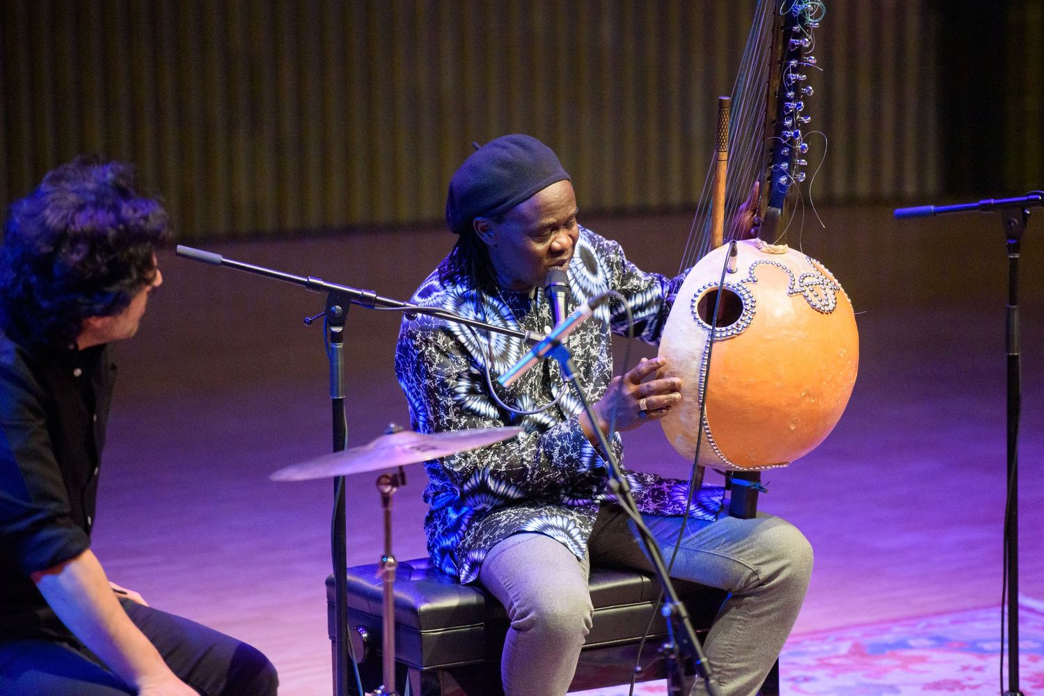 Musician playing a kora instrument on stage under purple lighting during a live performance.