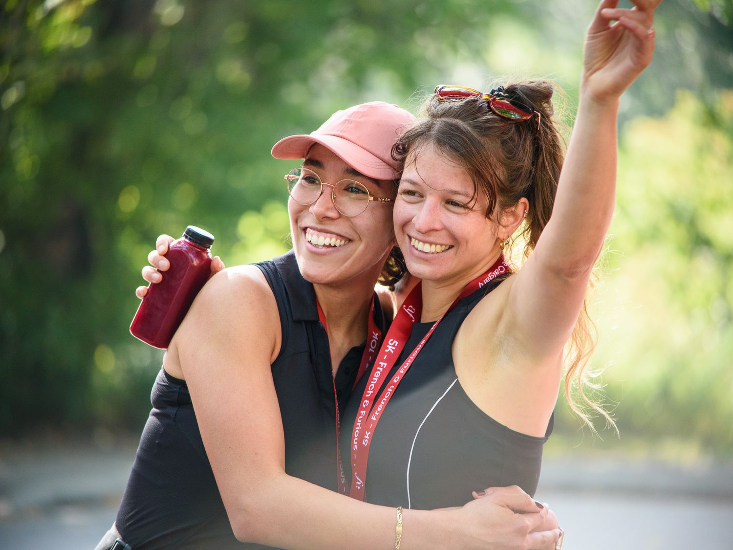 Two smiling women hugging outdoors, one wearing a pink cap holding a drink, the other with sunglasses on her head.