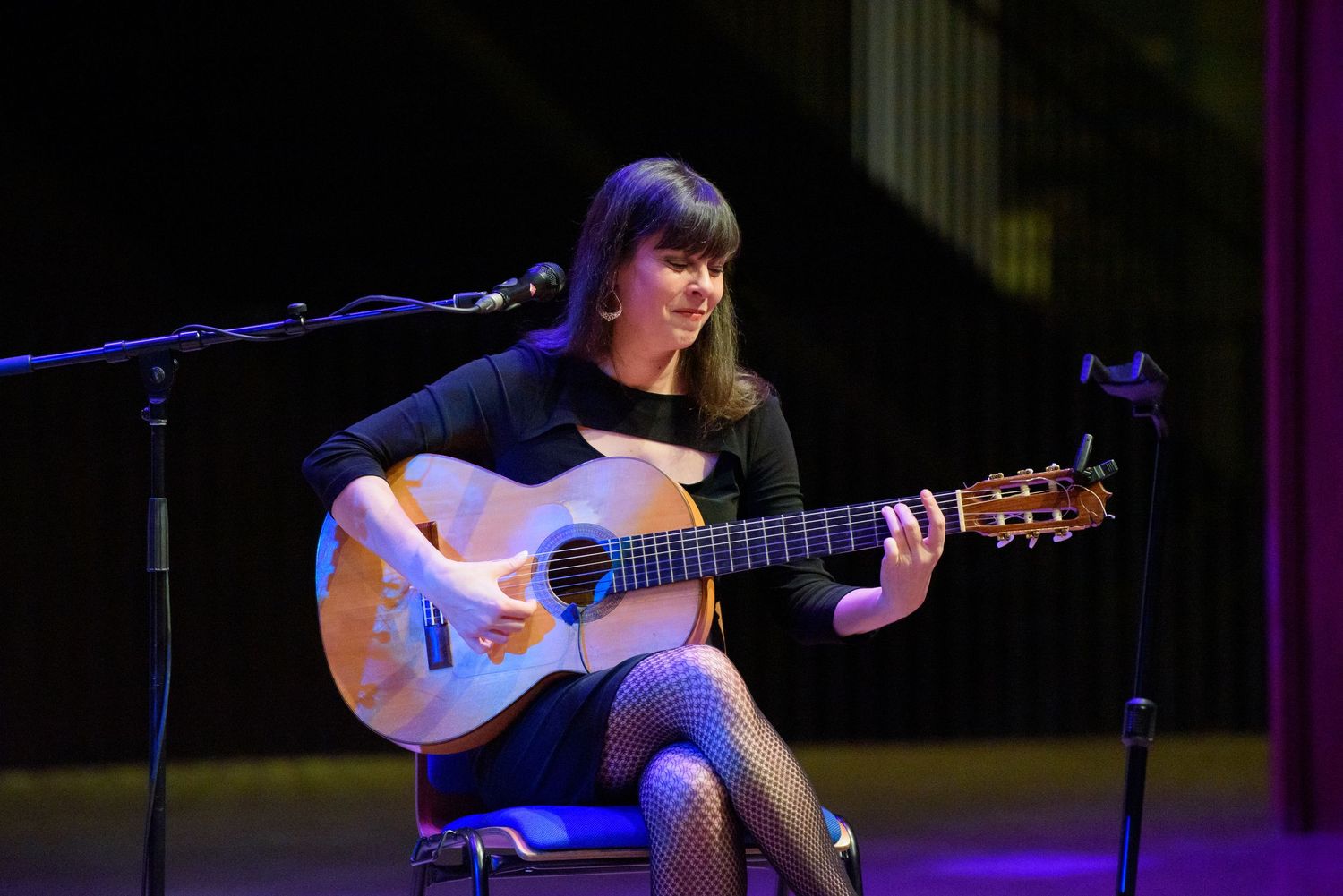 Young woman in black dress playing acoustic guitar on stage near microphone under colorful stage lights.