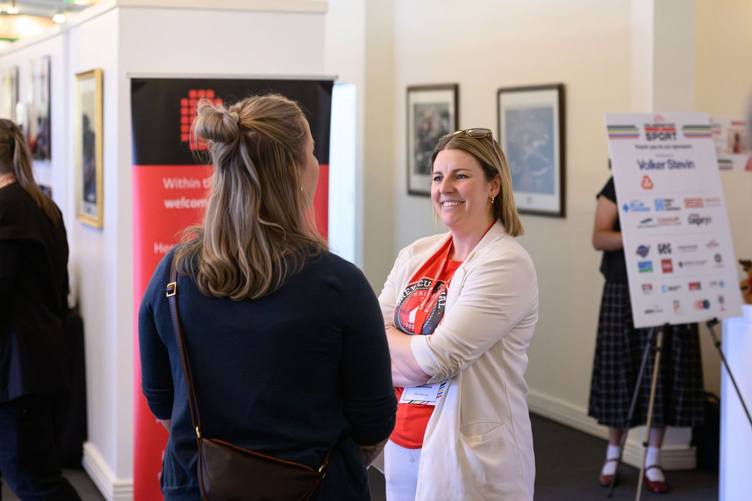 Two women smiling and chatting at an indoor networking event near a branded display board.