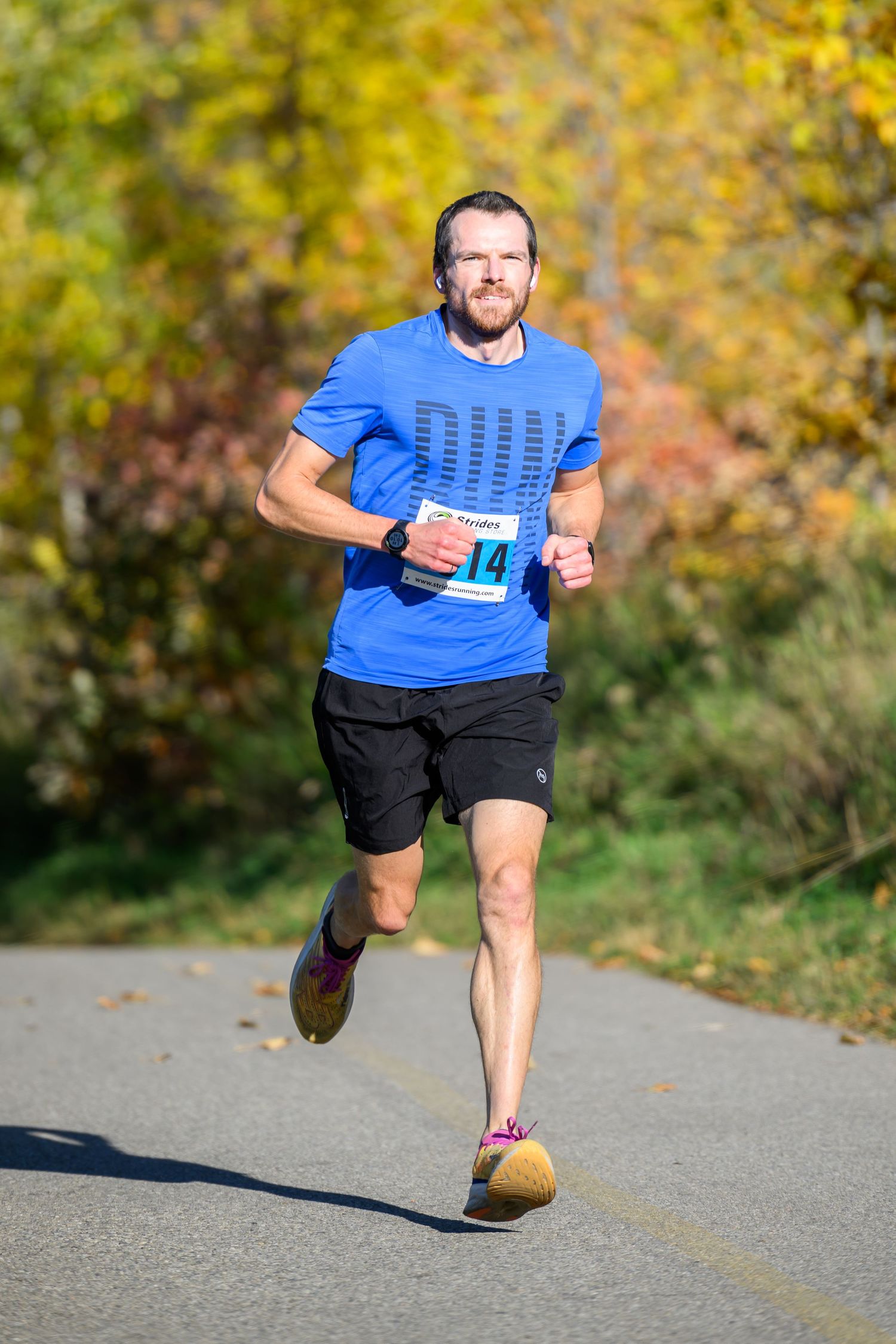 Male runner wearing bib number 14 races on a paved path surrounded by vibrant autumn foliage.