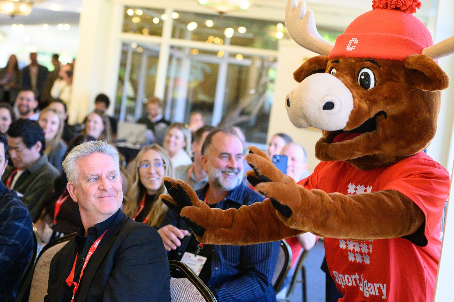 A moose mascot in a red Sport Calgary jersey poses with smiling attendees at an indoor event.