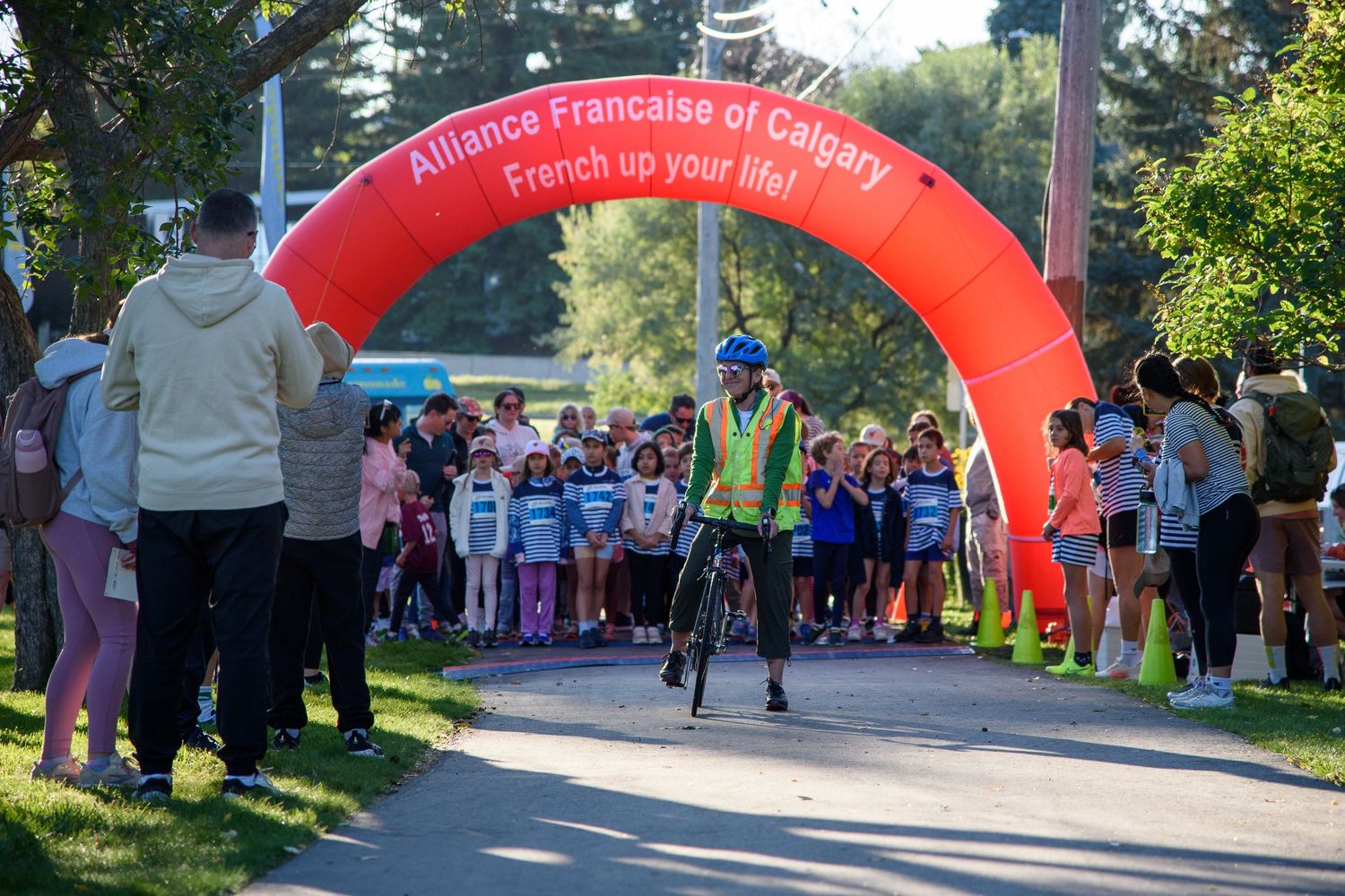 Cyclist rides under Alliance Francaise de Calgary inflatable arch at outdoor community fun run event.