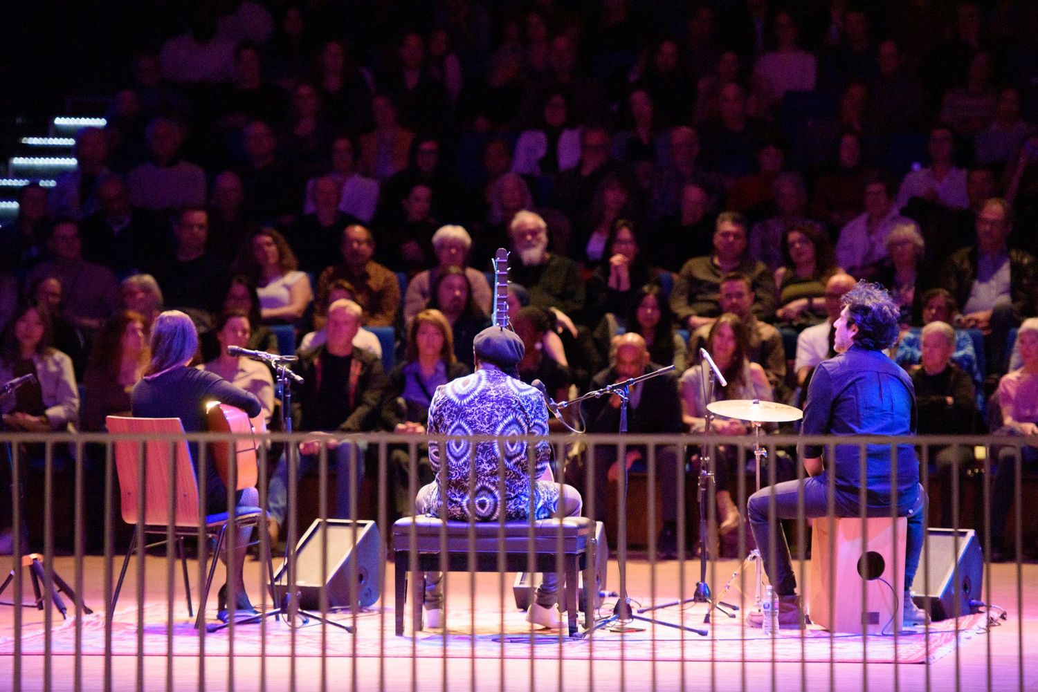 Musicians performing on stage in a concert hall surrounded by an attentive seated audience under dramatic lighting.