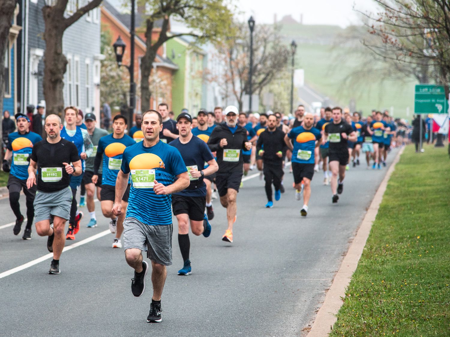Runners competing in an outdoor road race through a small town street, wearing blue shirts and race bibs on a cloudy day.