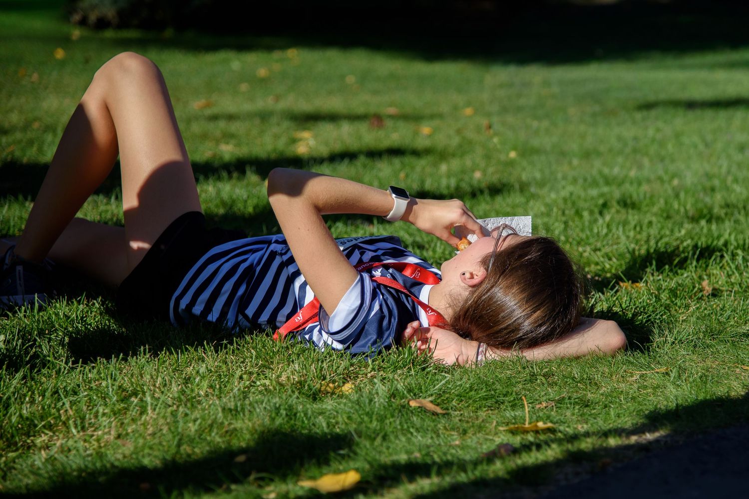 Young girl relaxing on green grass in sunny park, lying on her back in striped shirt on a bright summer day.