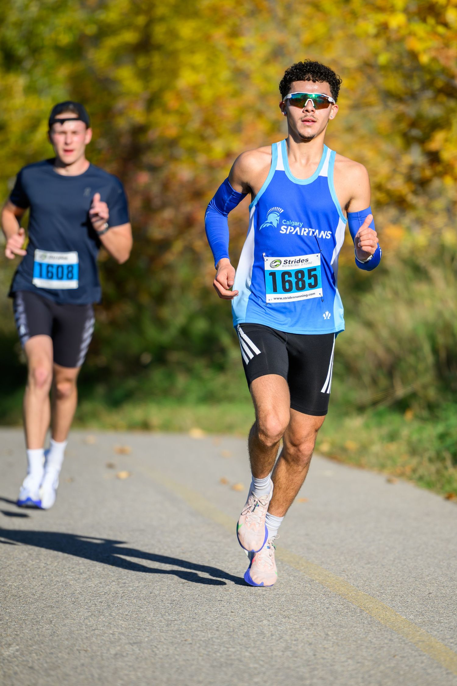 Two male runners competing in a fall road race, wearing numbered bibs 1685 and 1608, surrounded by autumn foliage.