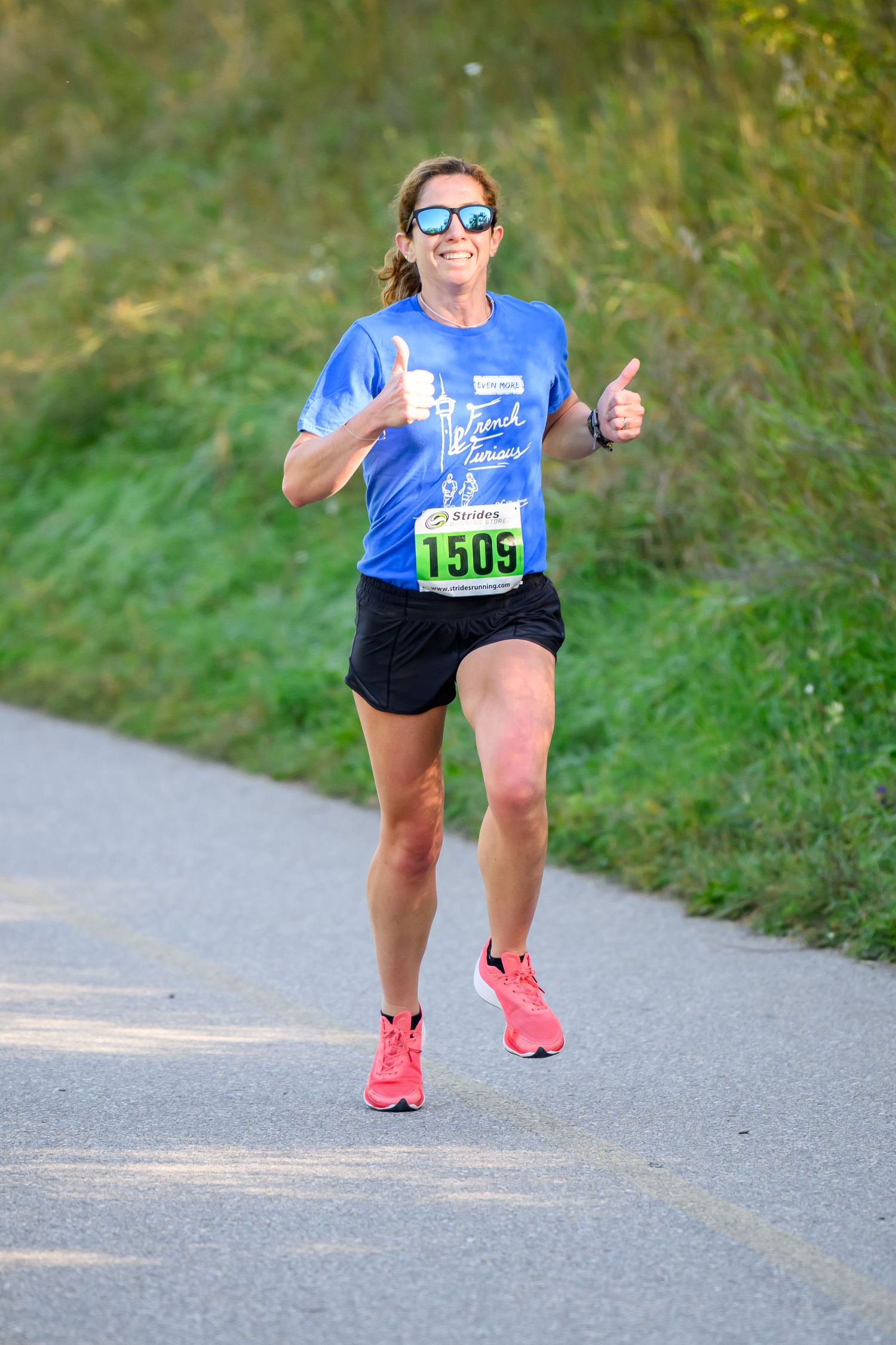 Smiling female runner #1505 giving thumbs up in blue shirt and pink shoes during outdoor road race.