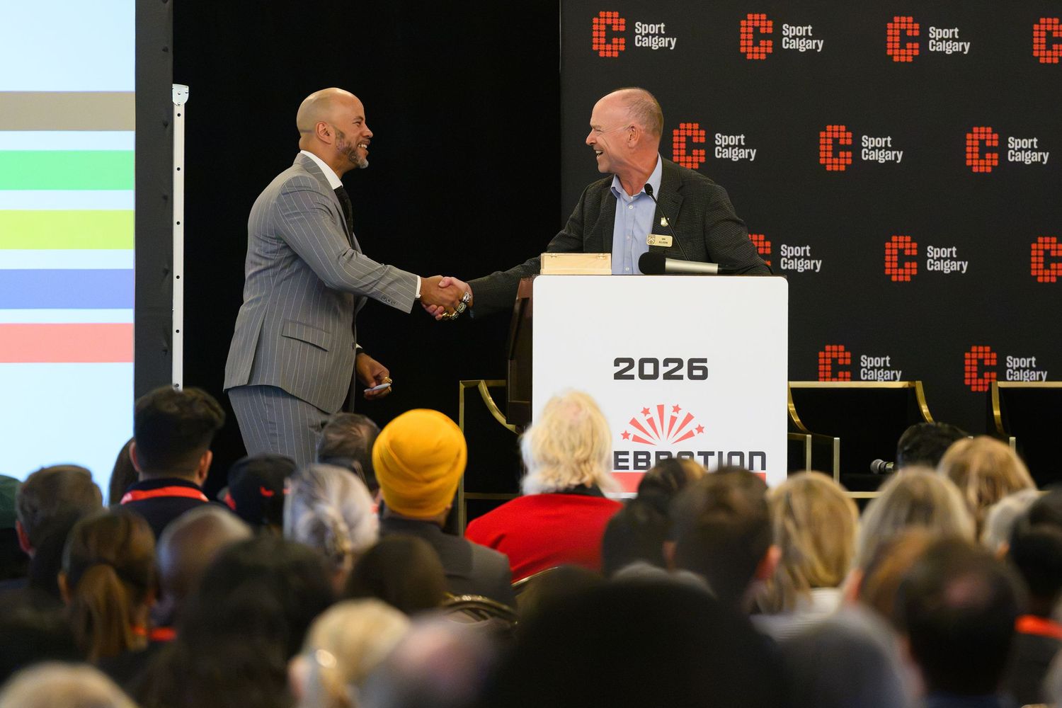 Two men shake hands on stage at a 2026 event in Calgary, with an audience seated in the foreground.