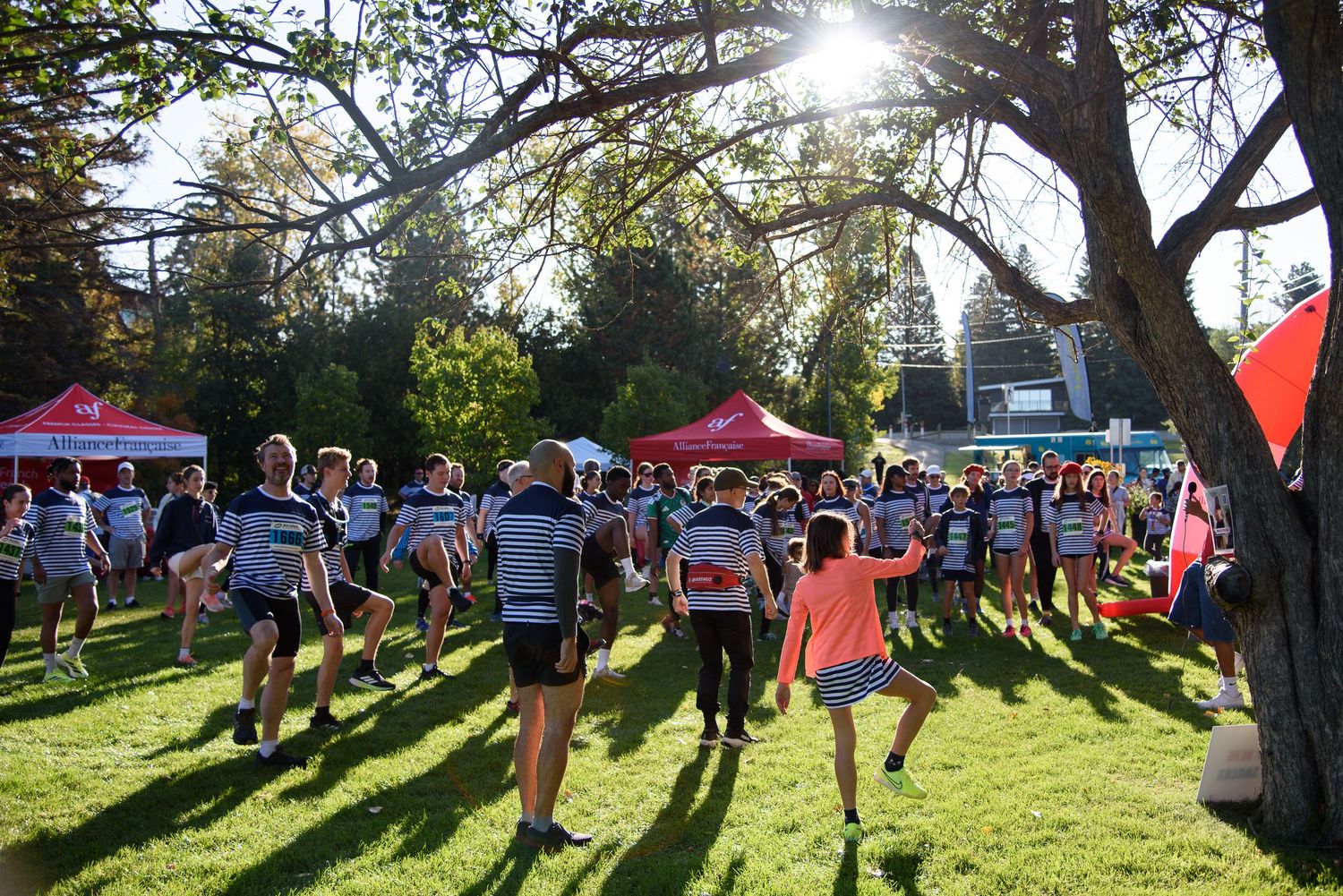 Crowd of rugby players and spectators gather on sunny grass field under trees near red event tents outdoors.