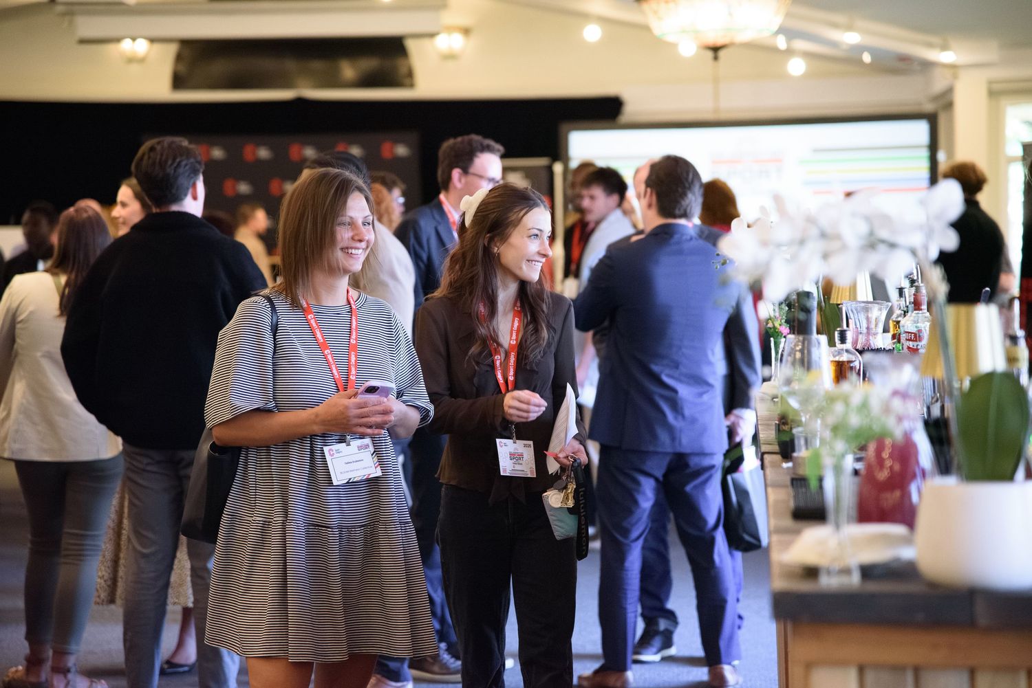 Two women wearing lanyards networking and smiling at an elegant indoor corporate reception.