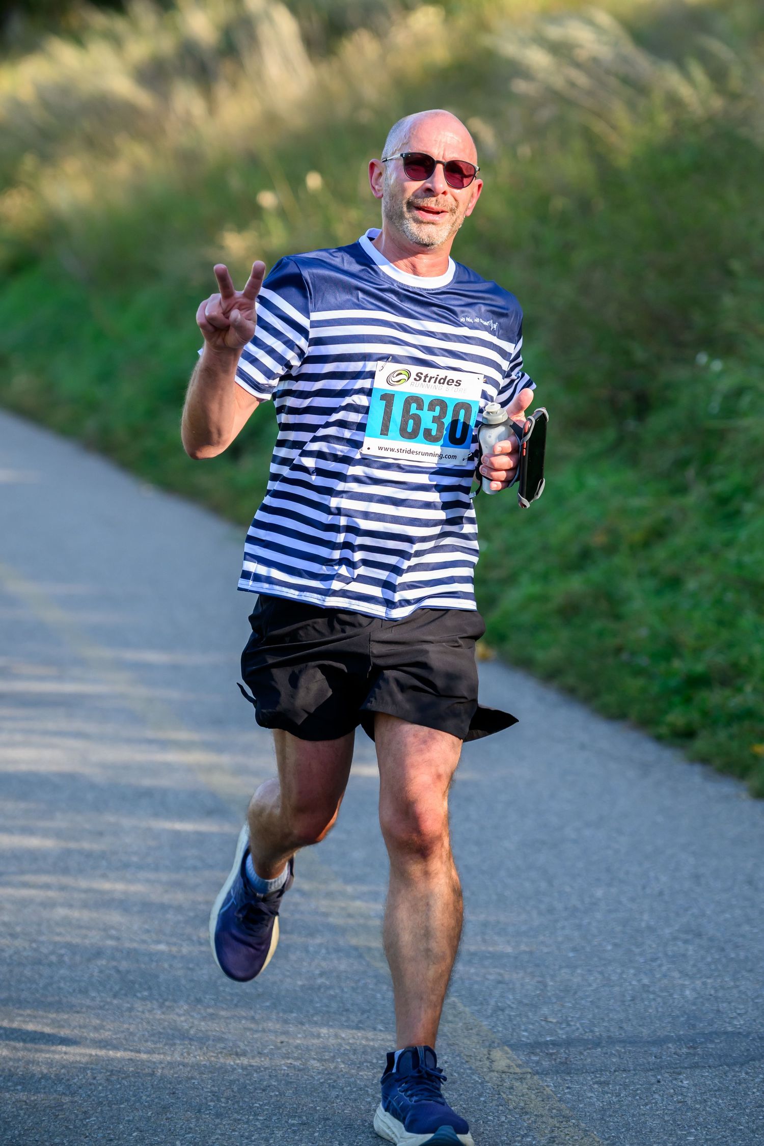 Bald male runner wearing bib number 1630 flashing peace sign during outdoor road race in striped shirt.