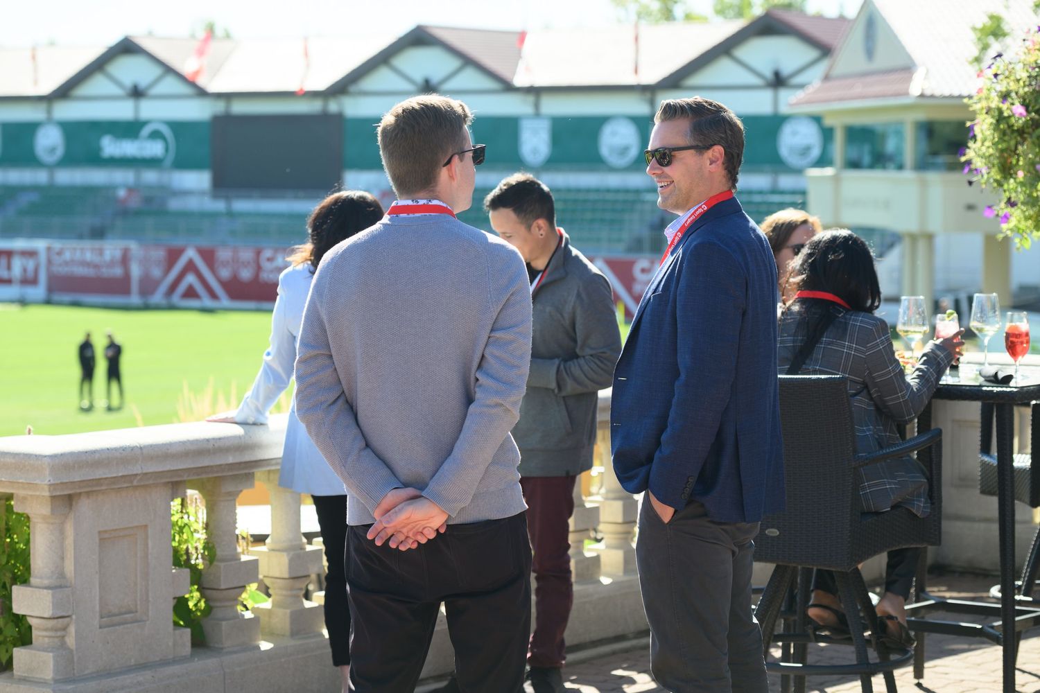 Men networking outdoors at a sports stadium venue on a sunny day, with a green football pitch visible in the background.