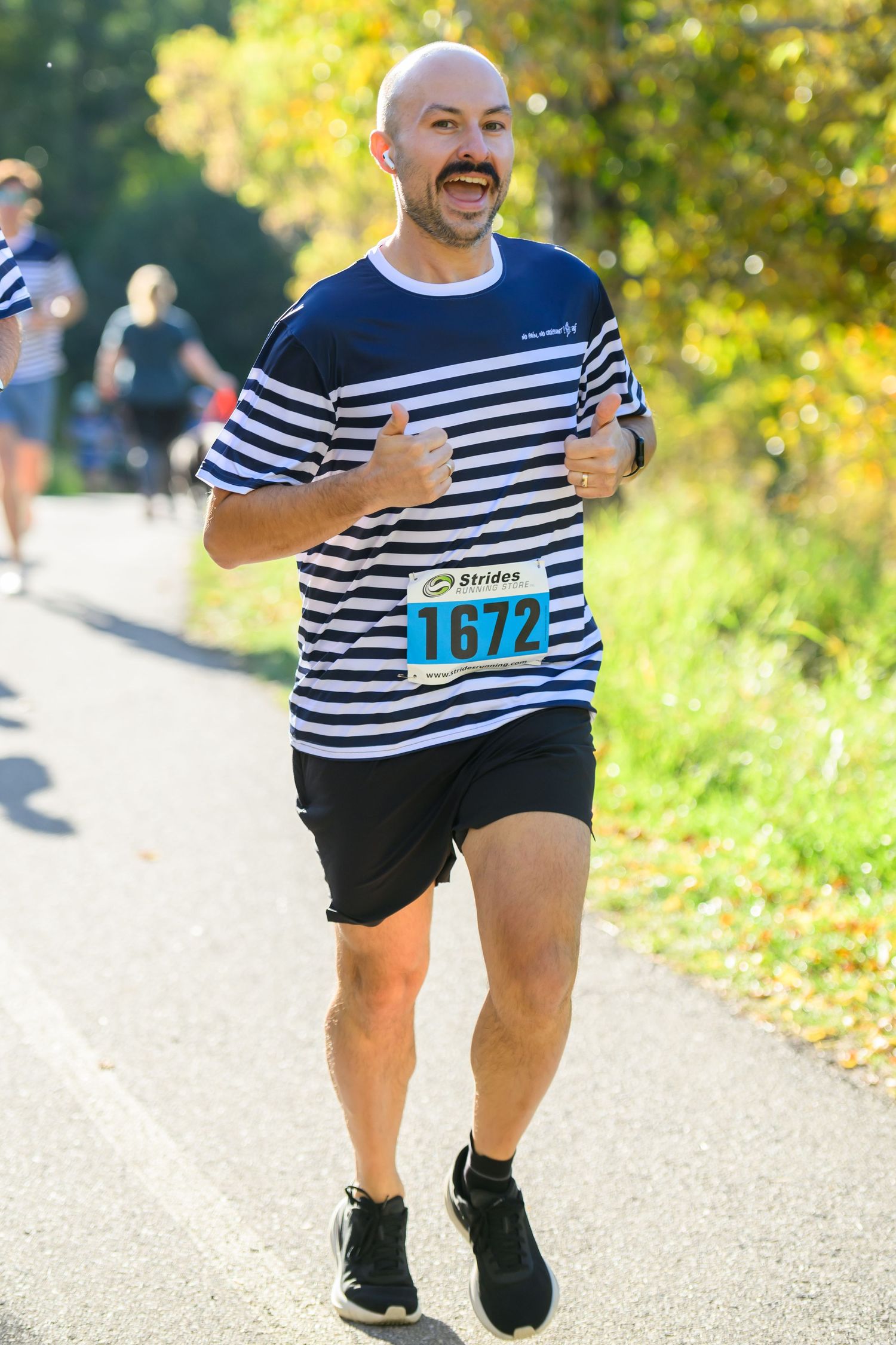 Bald man wearing striped shirt and race bib 1672 running in outdoor road race on sunny day.