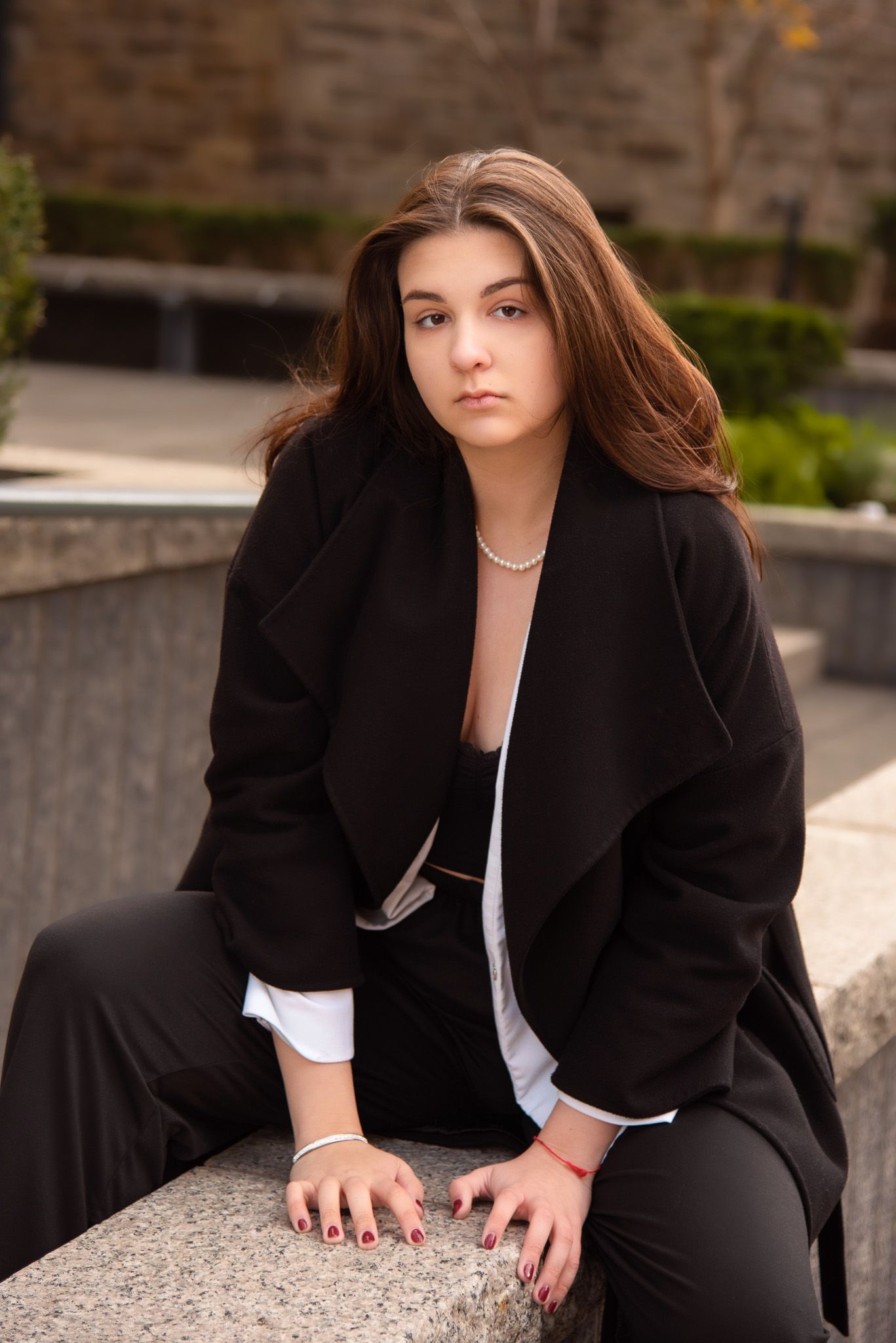 Young brunette woman in black blazer sitting on stone ledge in outdoor urban setting.