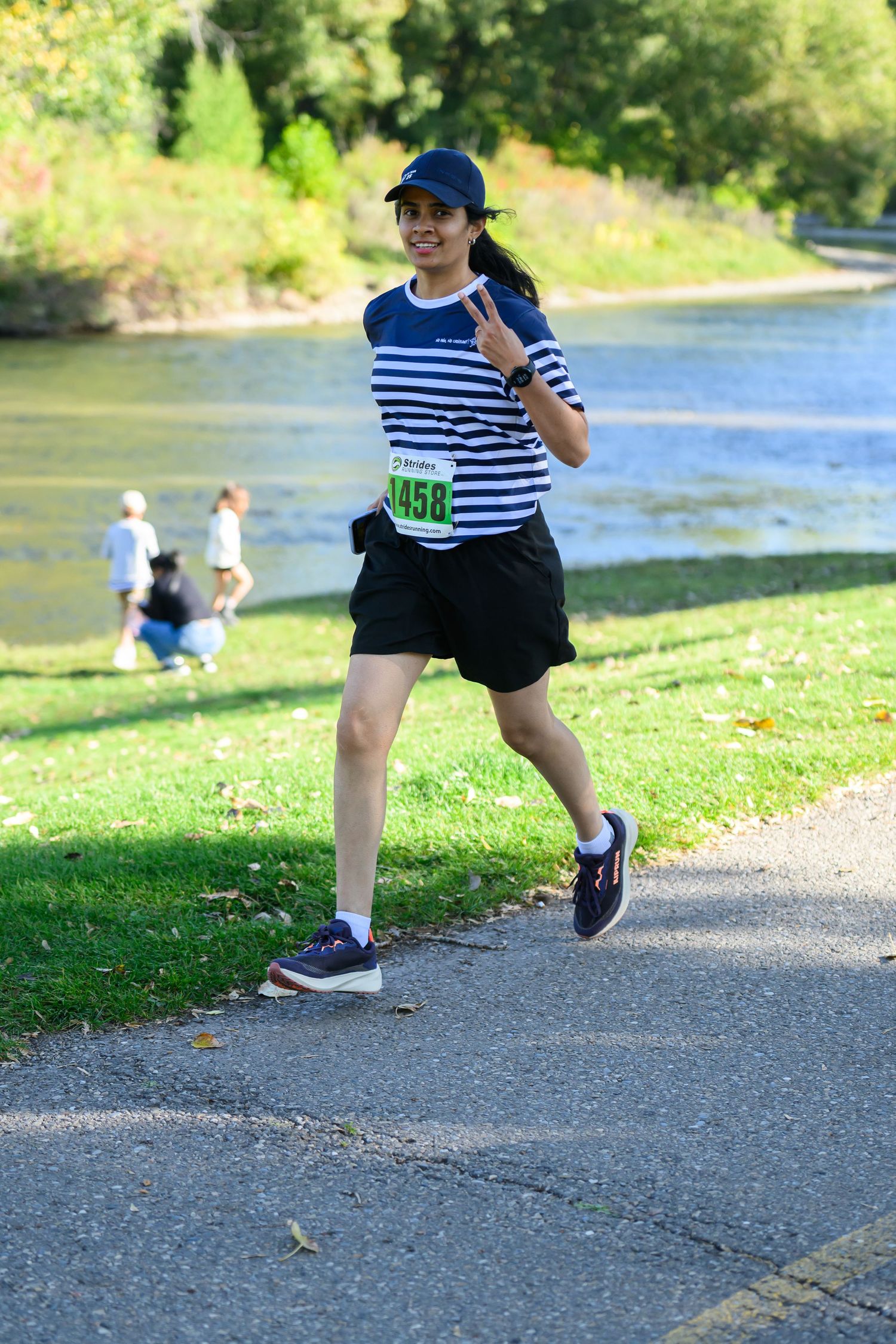 Smiling woman runner wearing bib 1458 flashing peace sign while jogging along a scenic riverside race course.