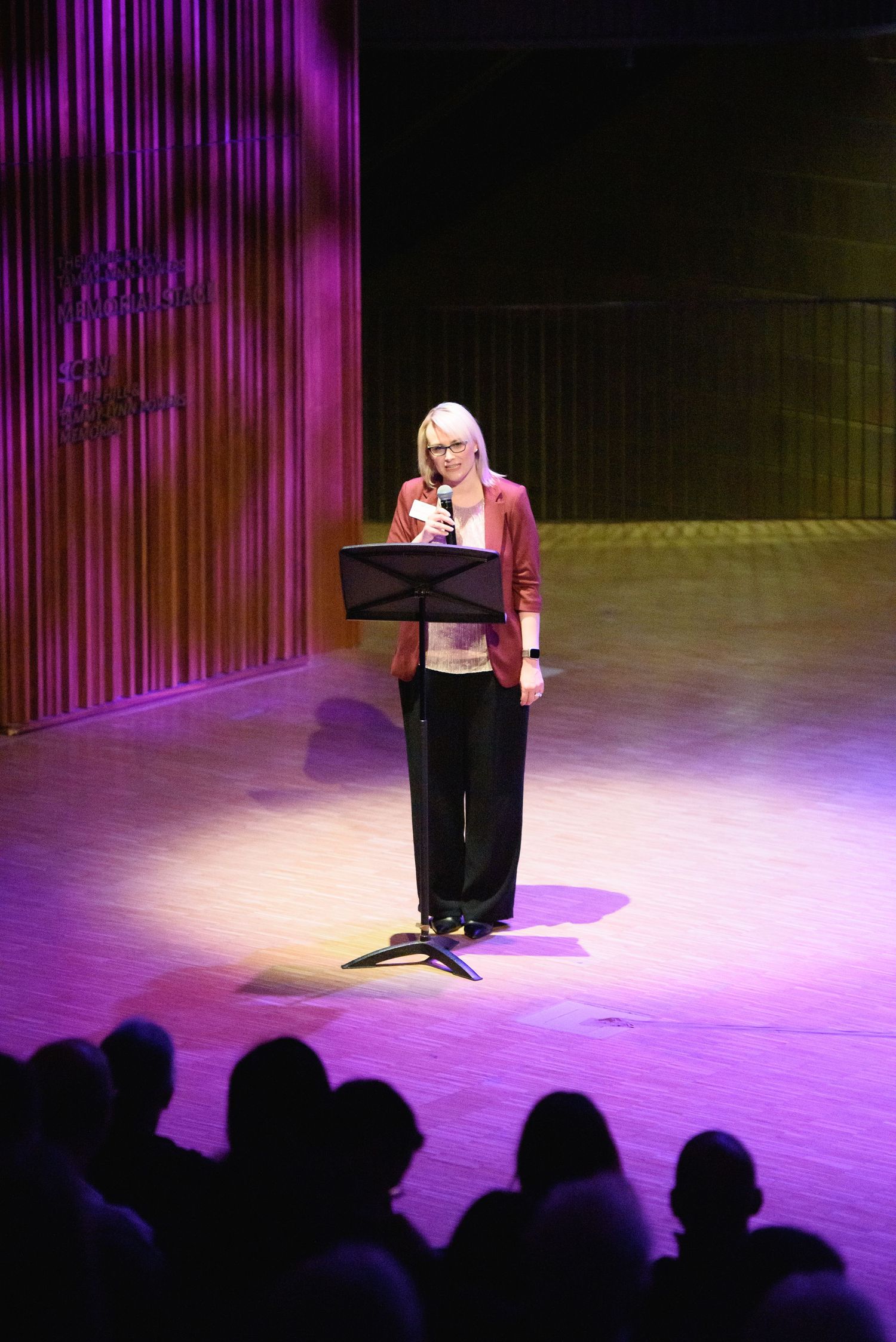 Woman in red blazer speaking at podium on purple-lit stage before audience.