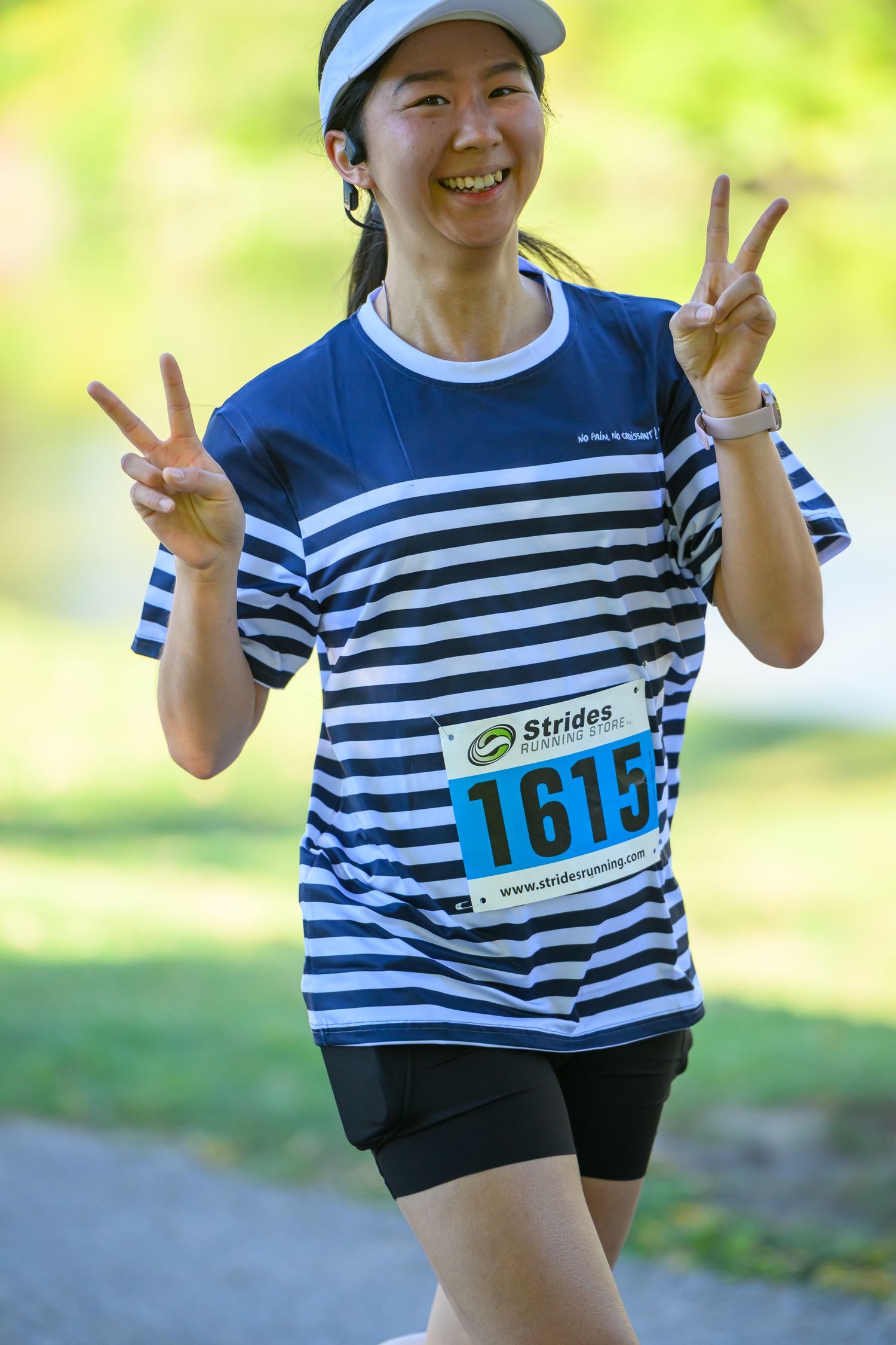 Smiling female runner #1615 flashing peace signs in blue striped shirt during Strides running race event.