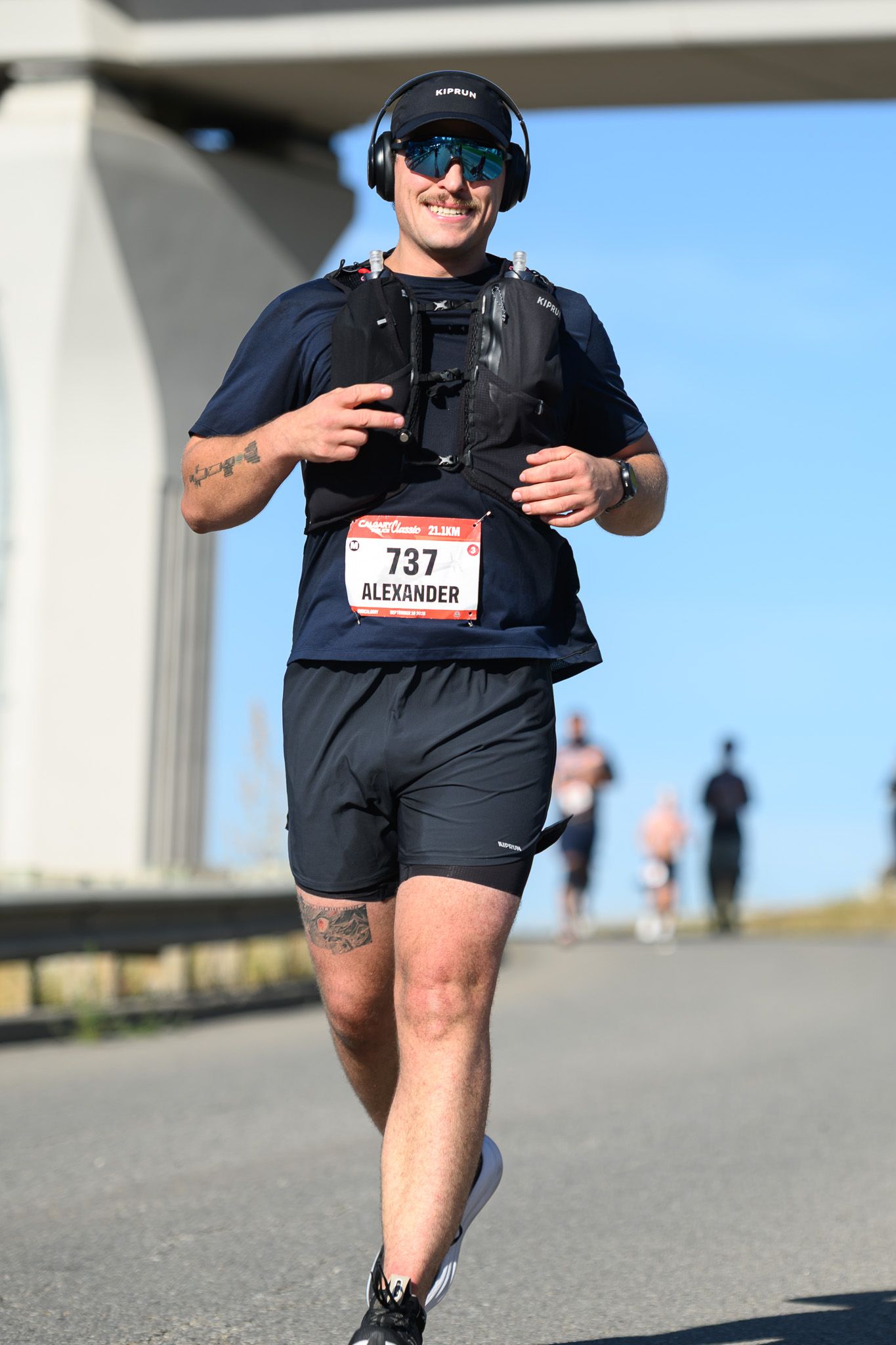 Runner Alexander, bib 737, wearing headphones and vest, smiles during a sunny road race.