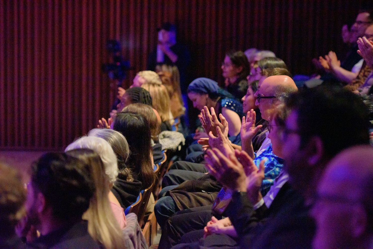 Engaged audience members enthusiastically clapping in a purple-lit theater during a live show.