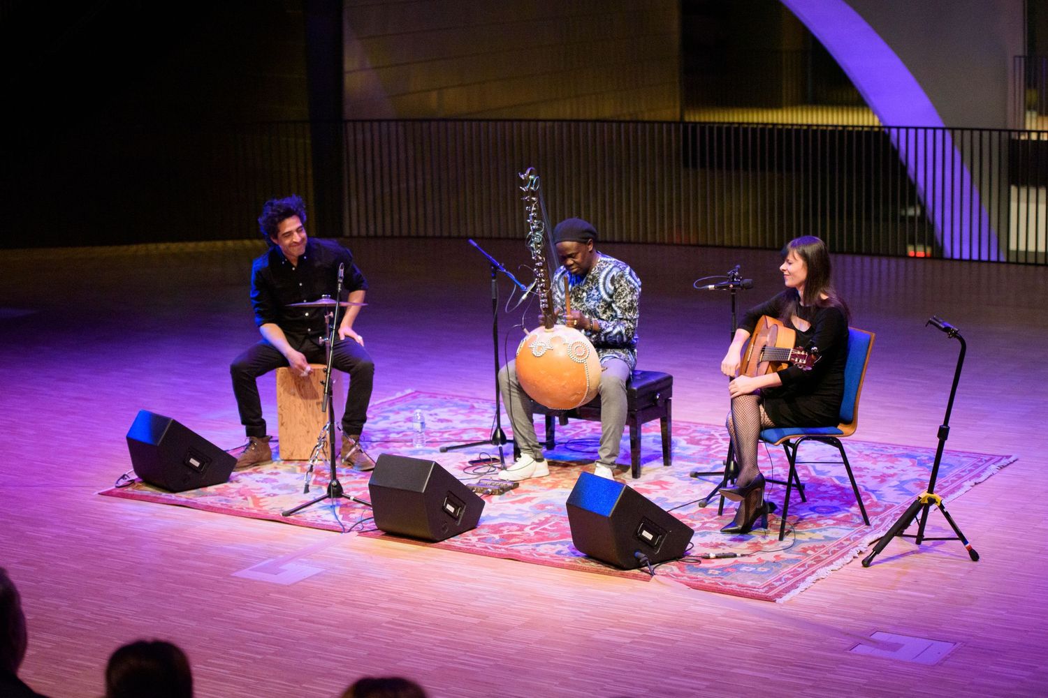 Three musicians perform world music on stage with traditional instruments under purple lighting at a concert venue.