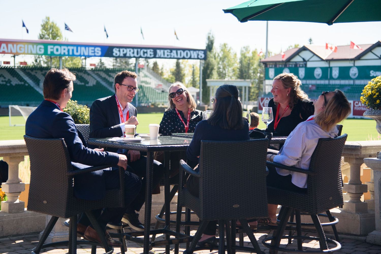 Six people sit around an outdoor patio table at Spruce Meadows equestrian venue on a sunny day.