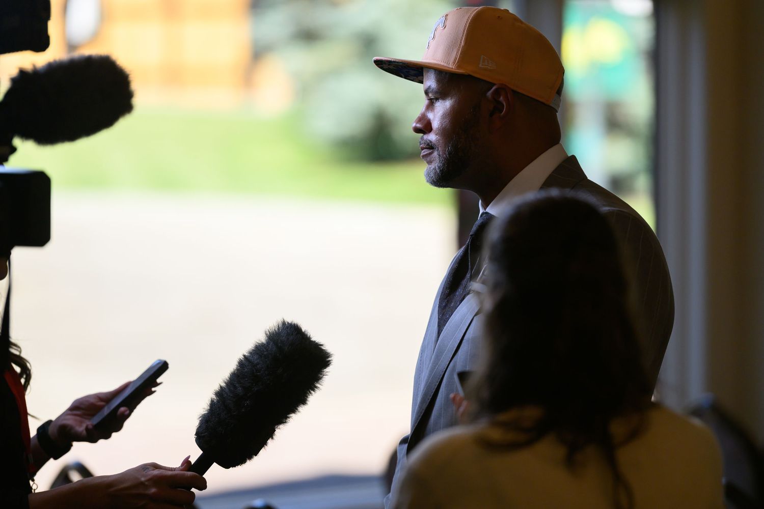 Man in yellow cap speaks to media reporters holding microphones at an outdoor press conference.