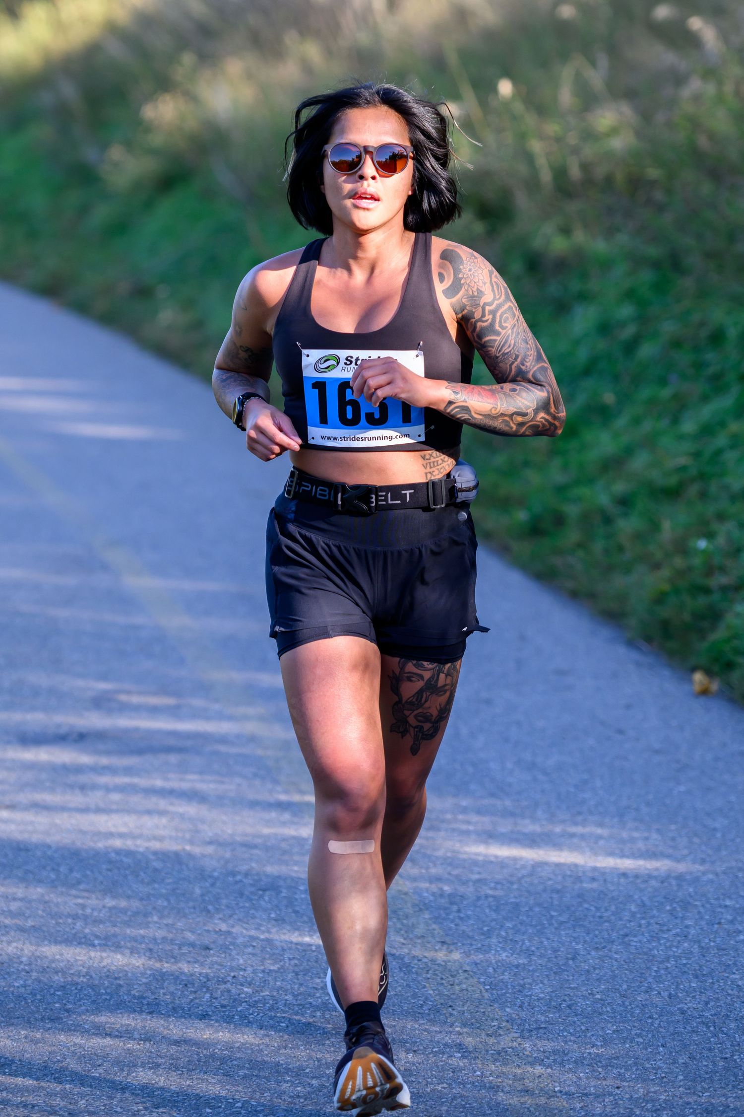 Tattooed female runner wearing bib number 1611 and sunglasses races on a paved path during an outdoor race.