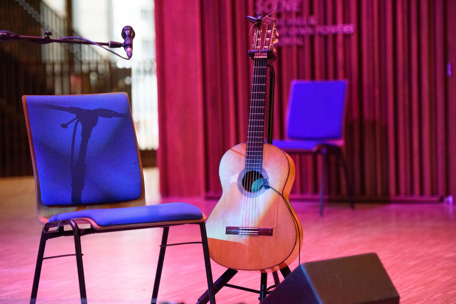 Acoustic guitar on a stand beside a music stand and chairs on a stage with red curtains and purple lighting.