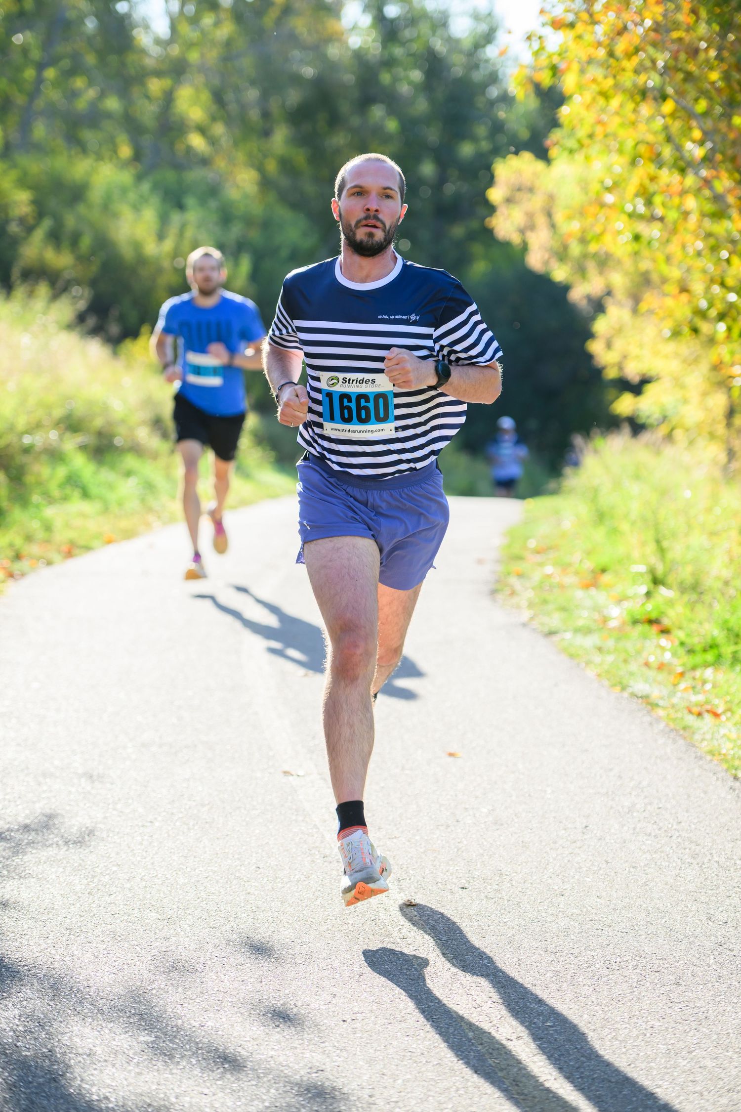 Bald male runner wearing bib number 1660 leads a road race on a sunny tree-lined path during an outdoor marathon event.