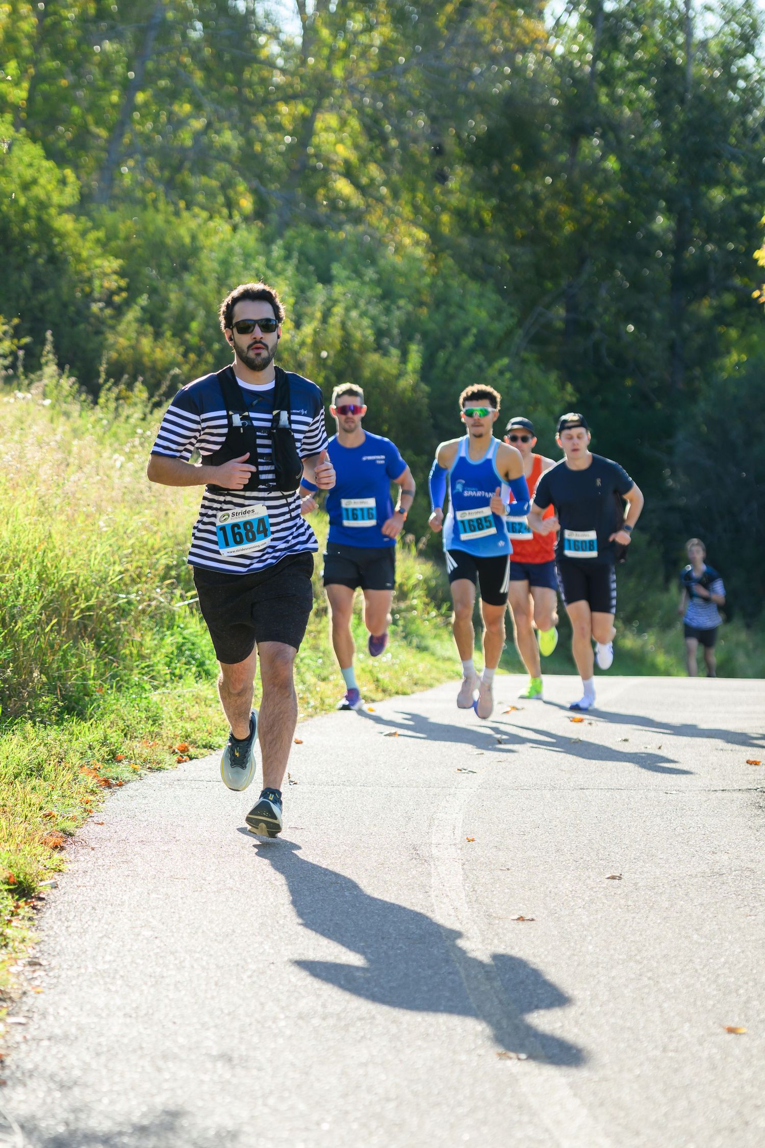 Runners compete in an outdoor road race on a sunny day, with lush green trees lining the path.