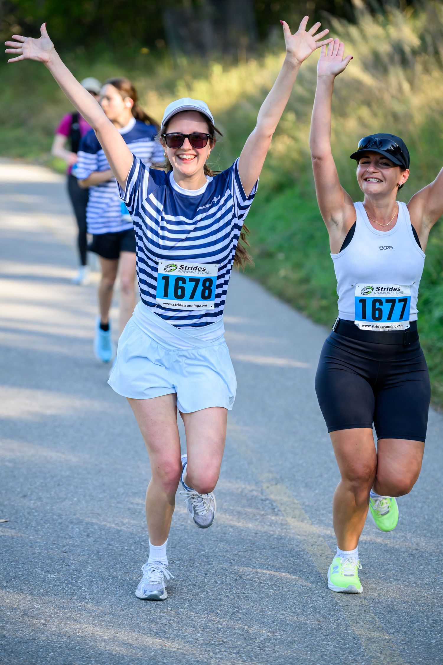 Two women runners celebrating with raised arms during a road race, wearing bib numbers 1677 and 1678.