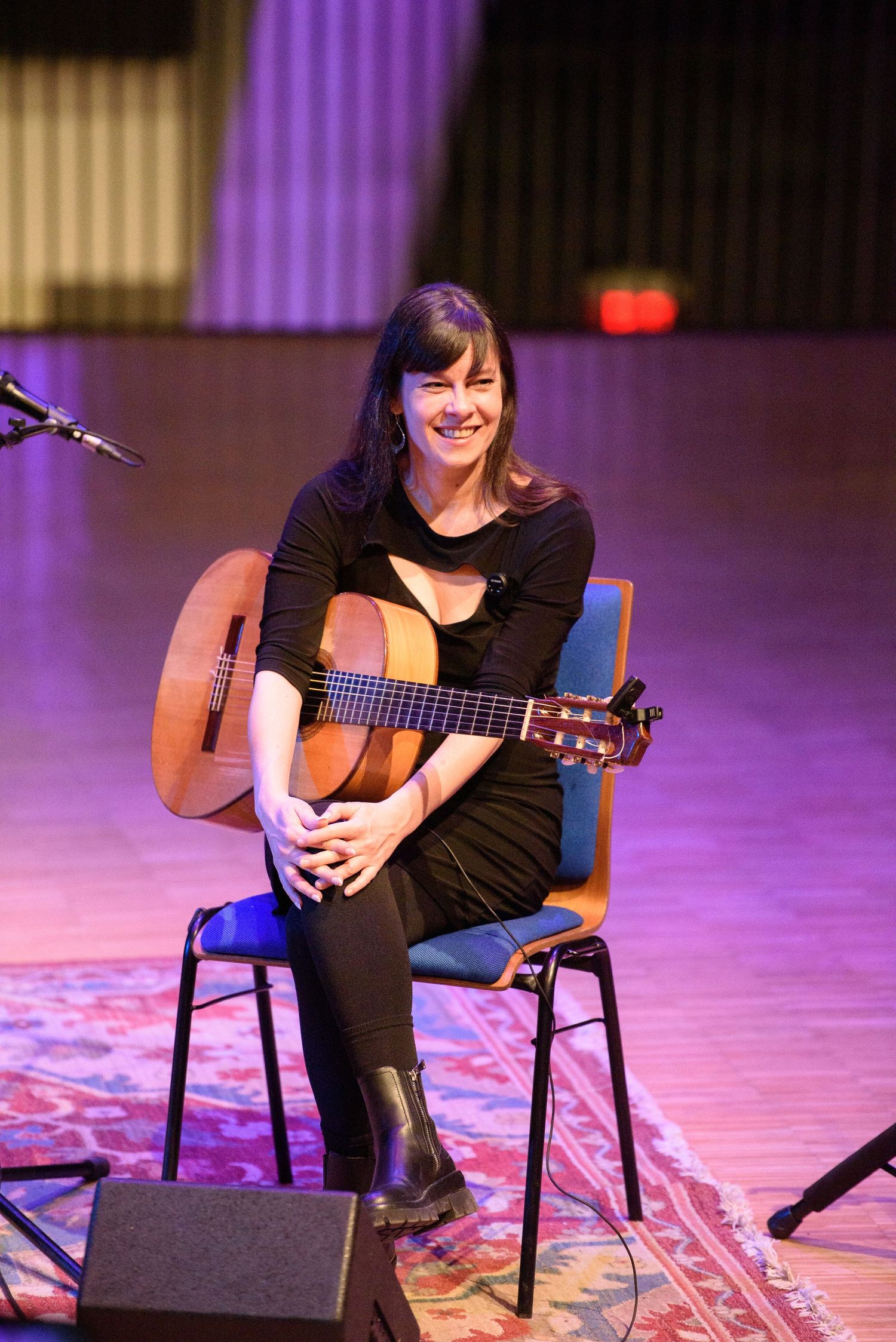 Smiling woman in black dress sitting on stage chair holding acoustic guitar during live music performance.