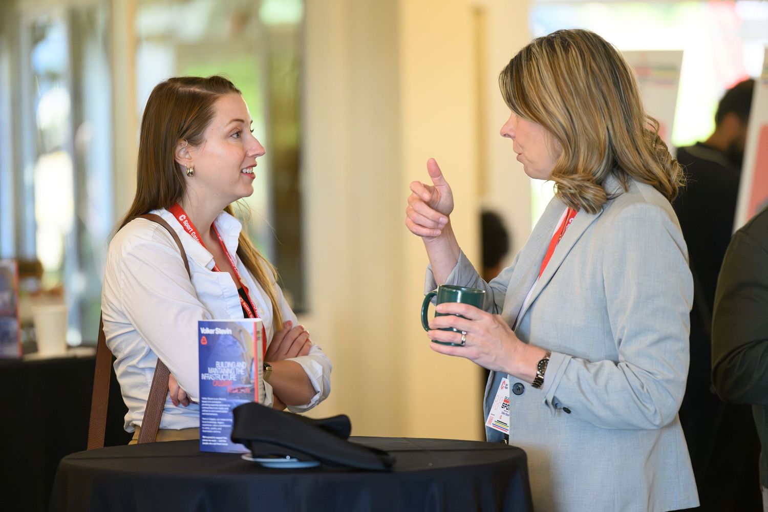 Two professional women networking at a conference, engaged in conversation near a round table with lanyards.