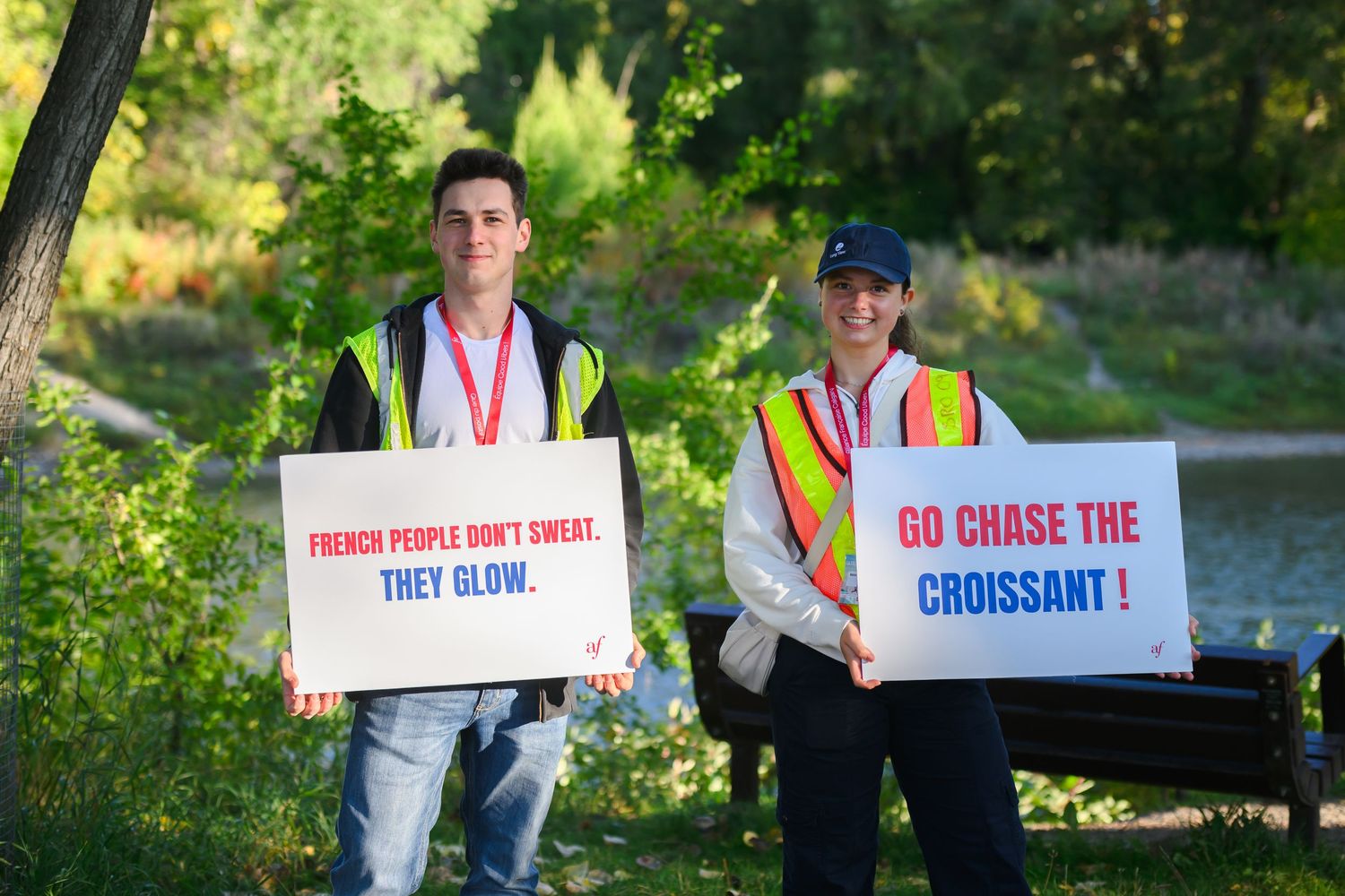 Two volunteers in safety vests hold humorous signs about French people and croissants at an outdoor event.