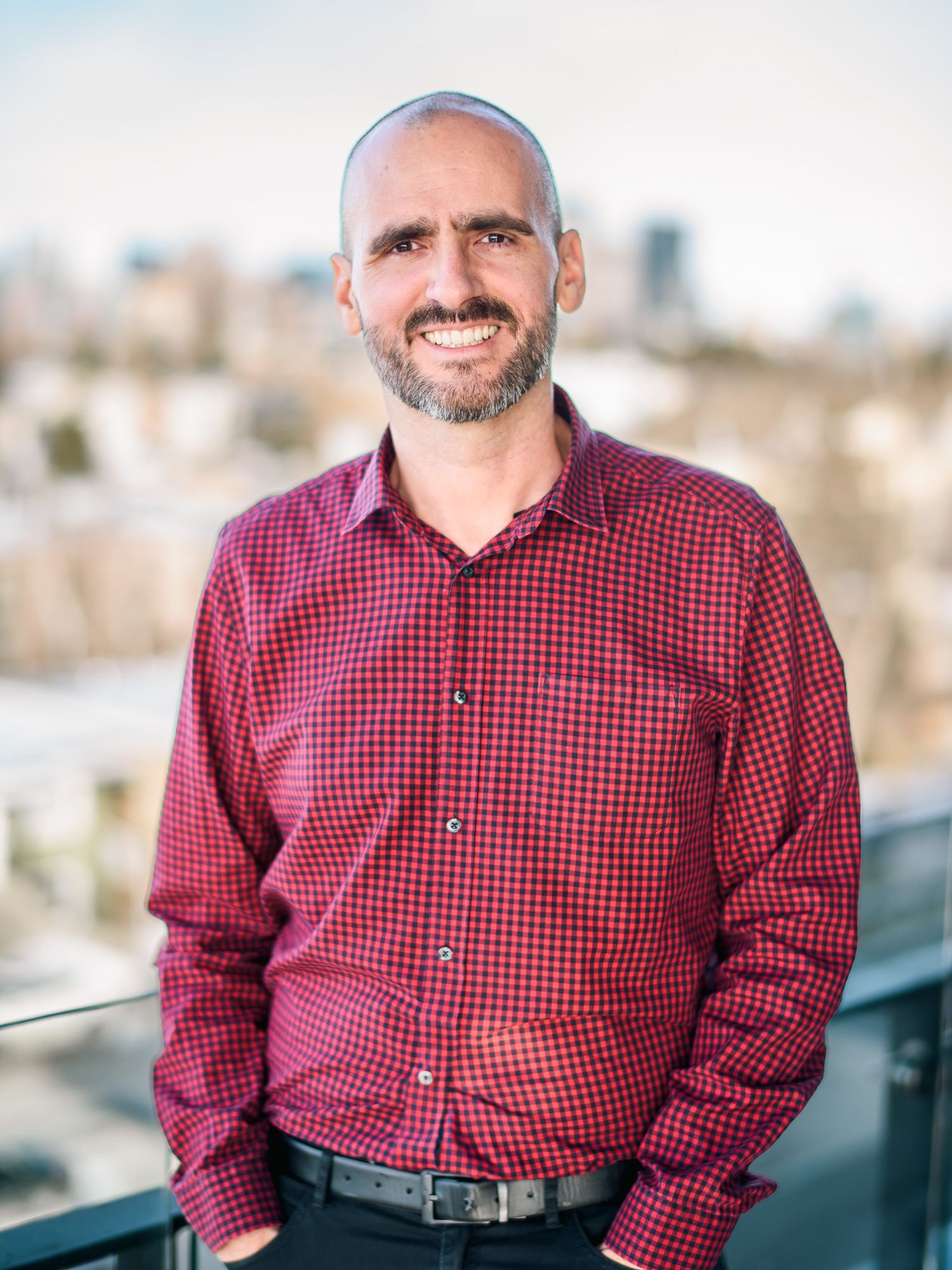Smiling middle-aged man in a red checkered shirt stands on an outdoor balcony with city views.