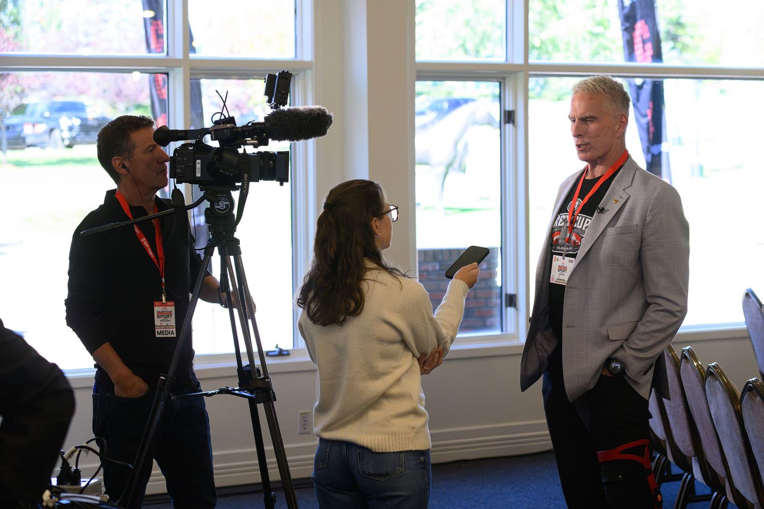 A TV camera crew films an interview with a tall man wearing a lanyard badge at an indoor event.