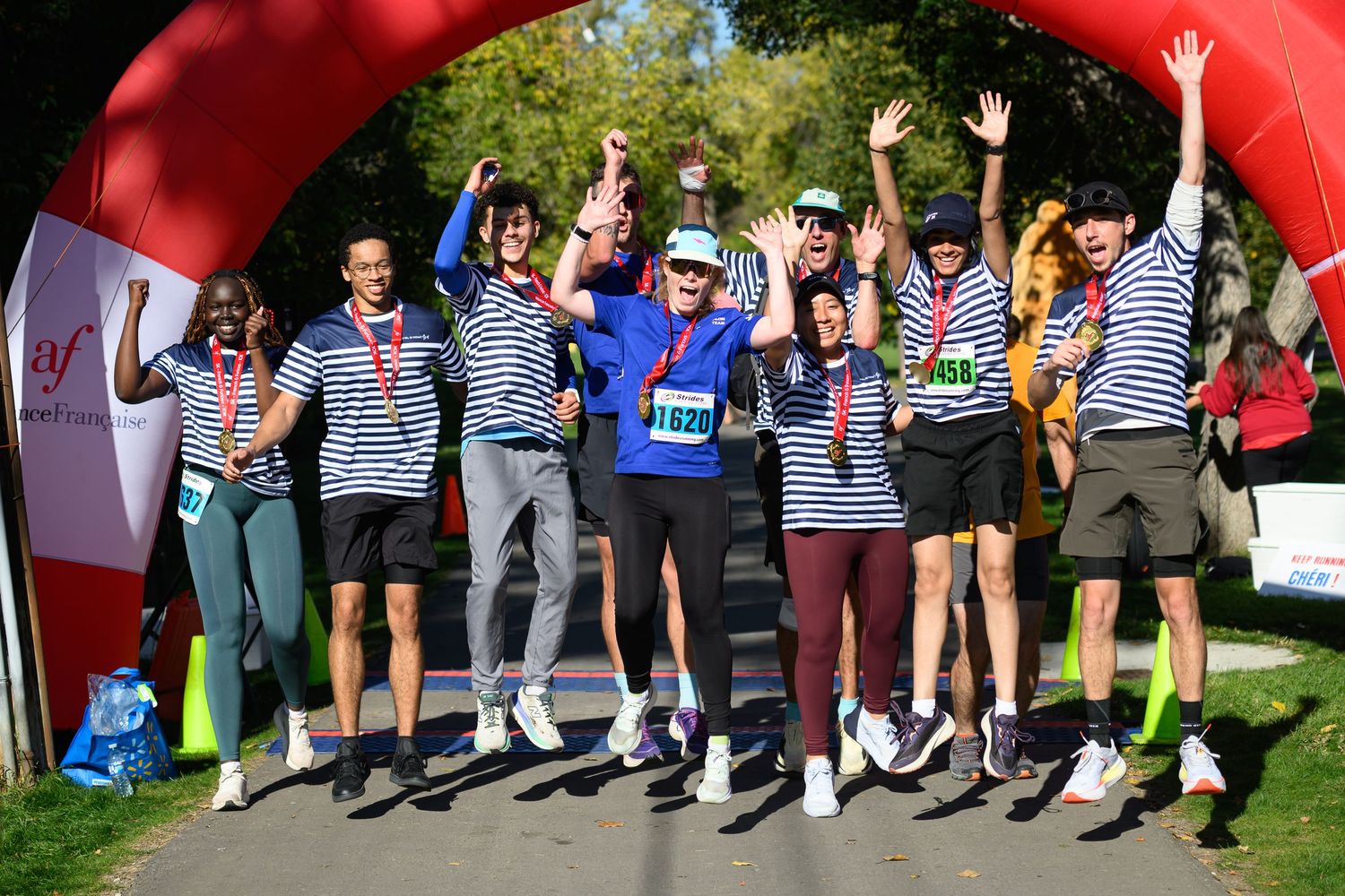 Group of runners celebrating at finish line arch, wearing matching striped shirts and race medals at outdoor charity run.