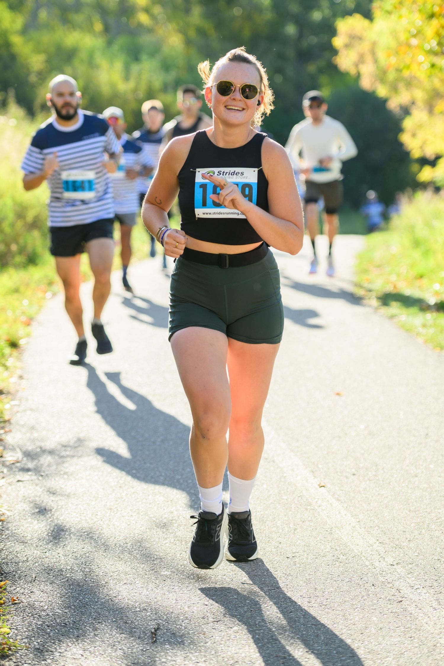 Smiling woman in black tank top and shorts runs a road race wearing bib number 1180 on a sunny tree-lined path.