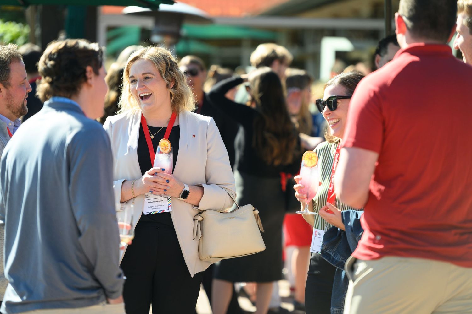 People socializing outdoors at a sunny event, woman in blazer laughing while holding a colorful drink.
