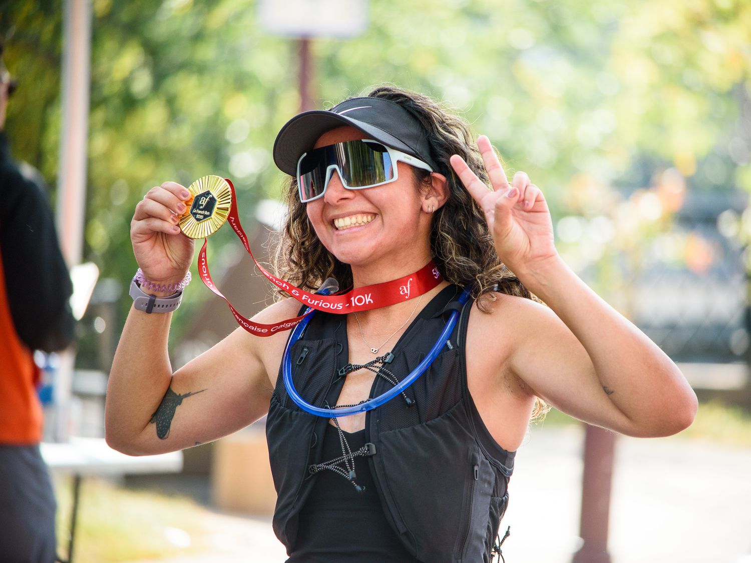 Smiling woman holding a gold finisher medal at a race event, wearing a black visor and sport sunglasses outdoors.