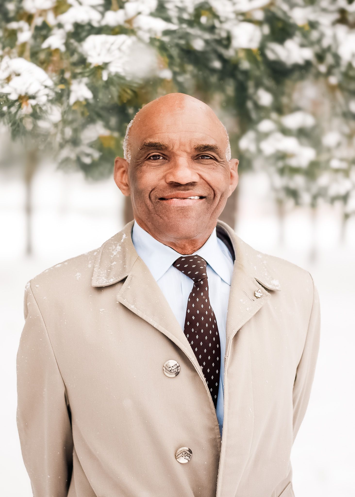 Smiling middle-aged Black man in beige trench coat and tie posing outdoors in snowy winter setting.