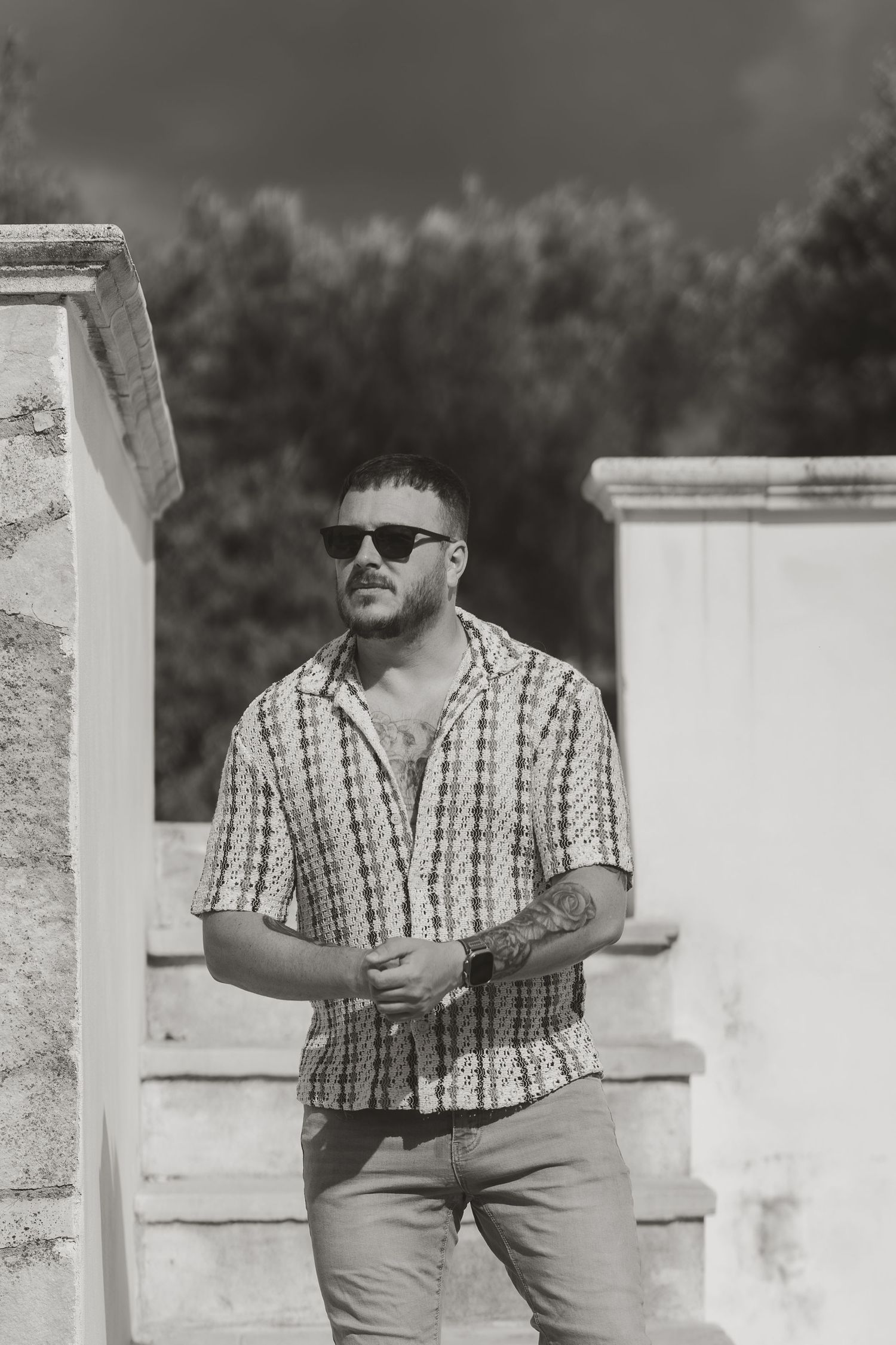 Tattooed man in patterned shirt and sunglasses standing by stone pillars outdoors.