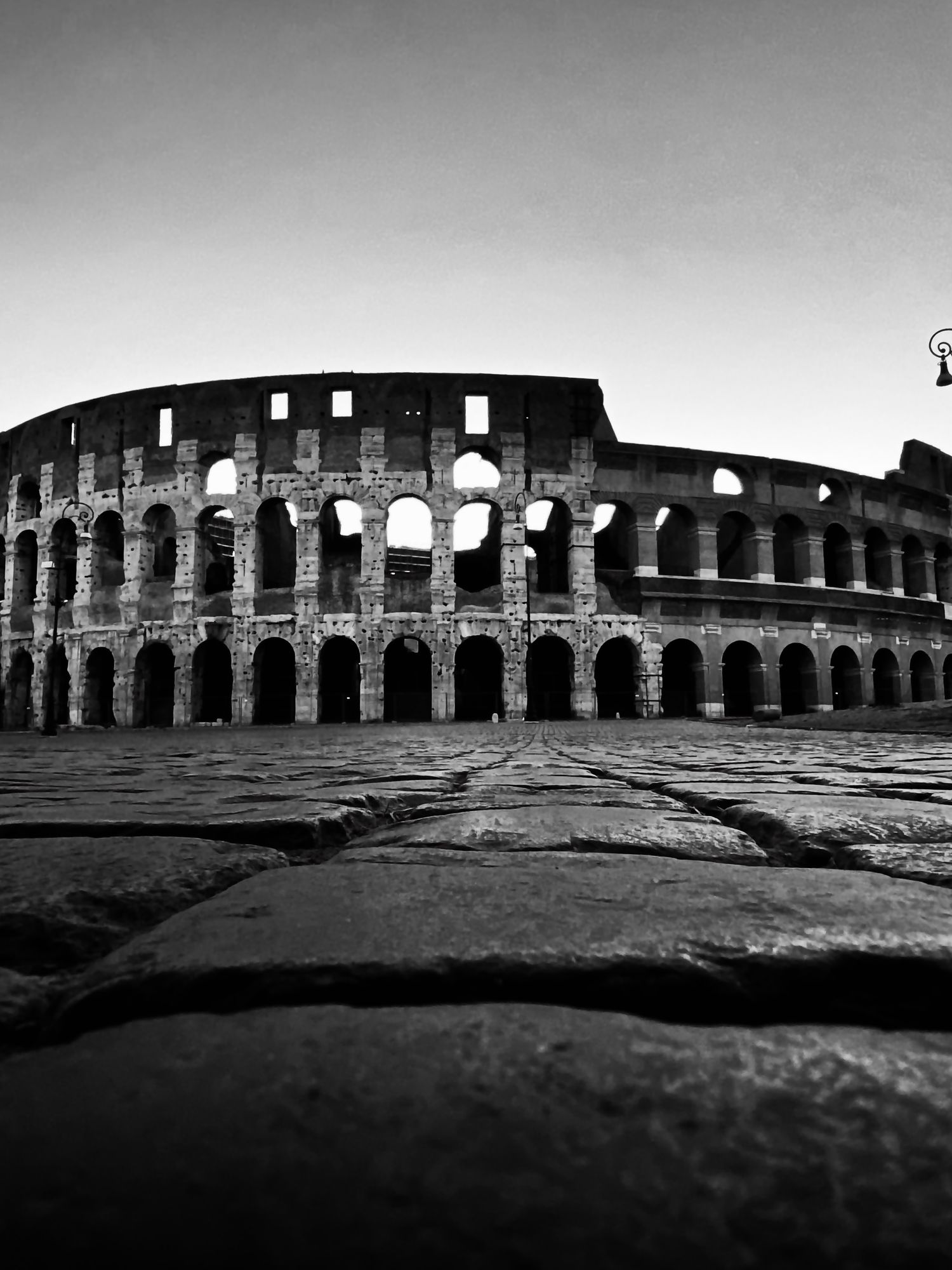 Low angle black and white photo of the ancient Roman Colosseum with cobblestone street foreground.