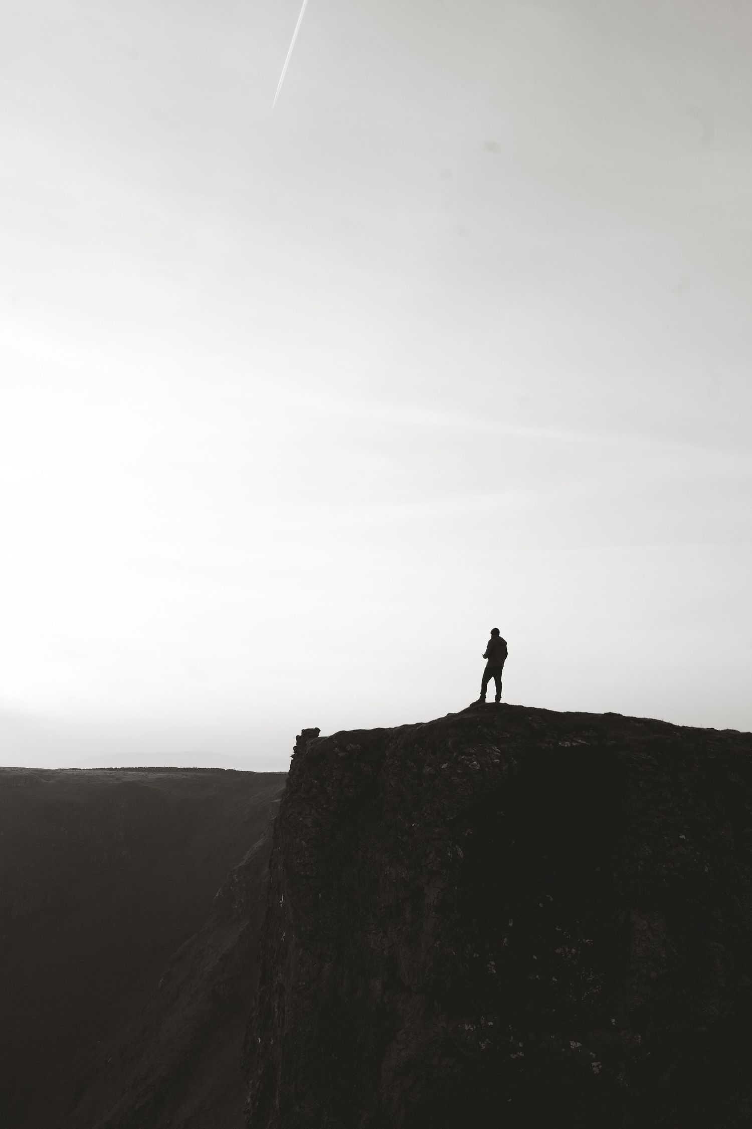 Silhouette of a lone hiker standing on a dramatic cliff edge against a bright overcast sky in black and white.