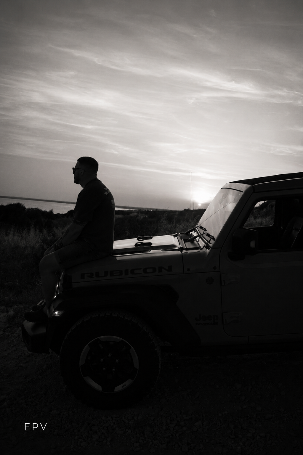Black and white silhouette of a man leaning on a Jeep Rubicon hood at sunset with scenic landscape behind.