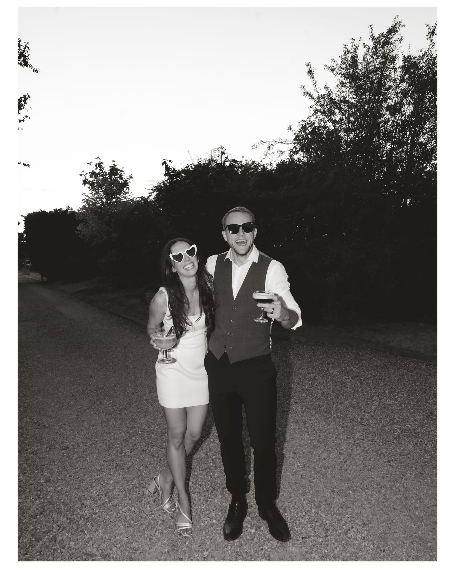 Couple in sunglasses holding drinks posing outdoors in black and white photo.
