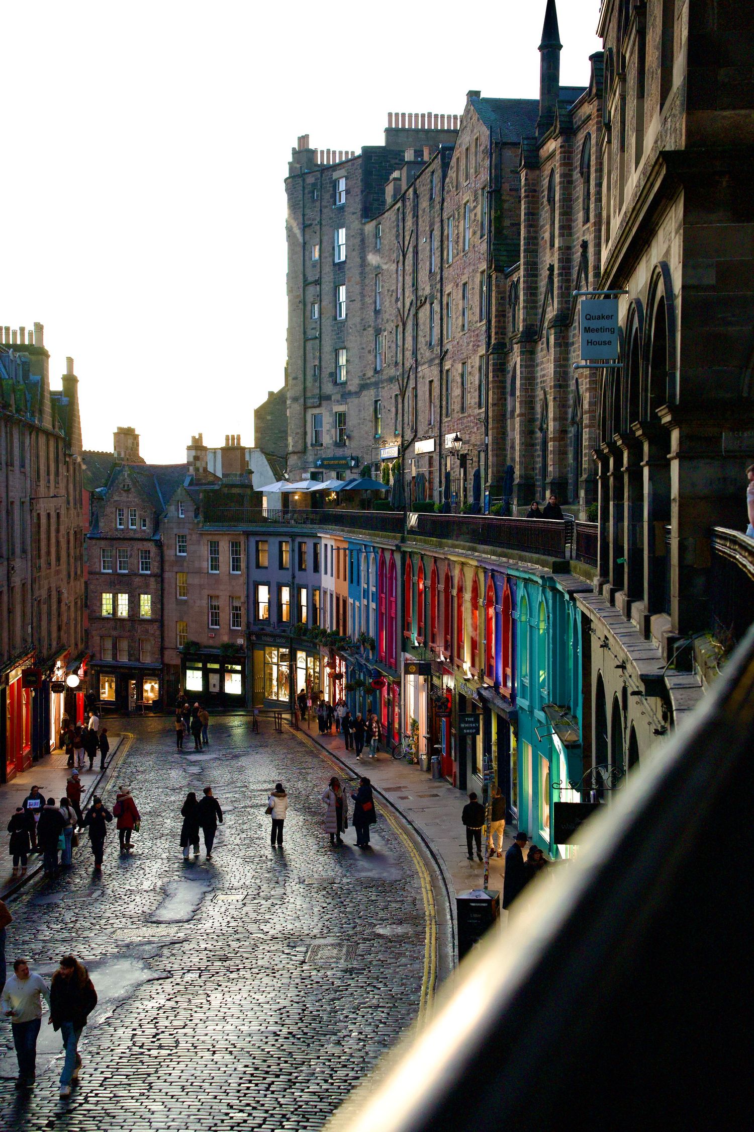Colorful shopfronts line the curved cobblestone Victoria Street in Edinburgh at golden hour with pedestrians.