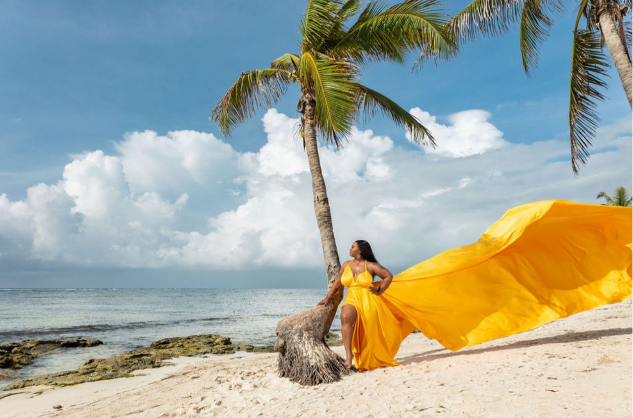 Woman in stunning yellow flowing dress leaning against palm tree on beach.