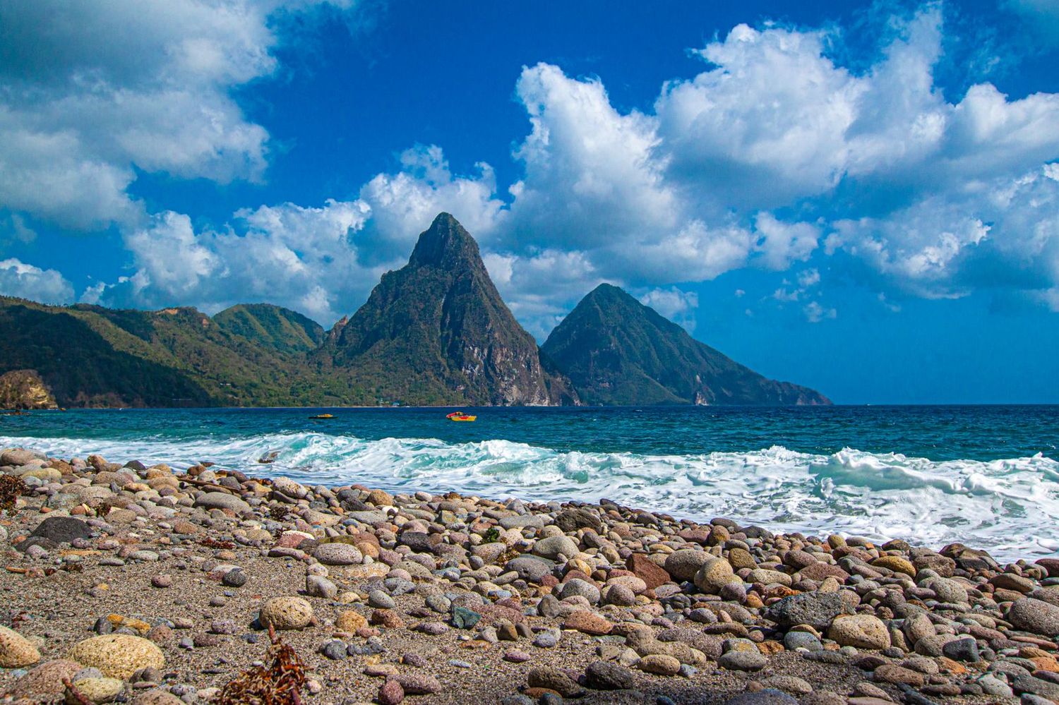 Rocky shoreline with turquoise waves and iconic twin Piton peaks rising dramatically under blue skies in Saint Lucia.